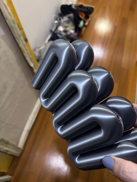Hand holding a glossy gray serpentine radiator-style object with rounded zigzag loops over a polished hardwood floor.