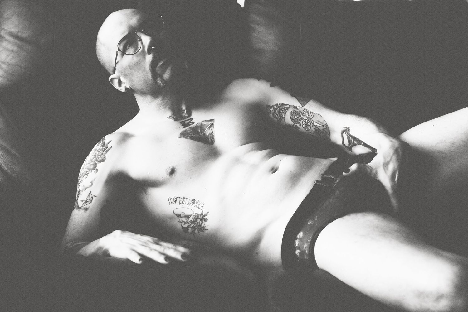 Black and white boudoir photo of a tattooed bald man reclining on a leather sofa.