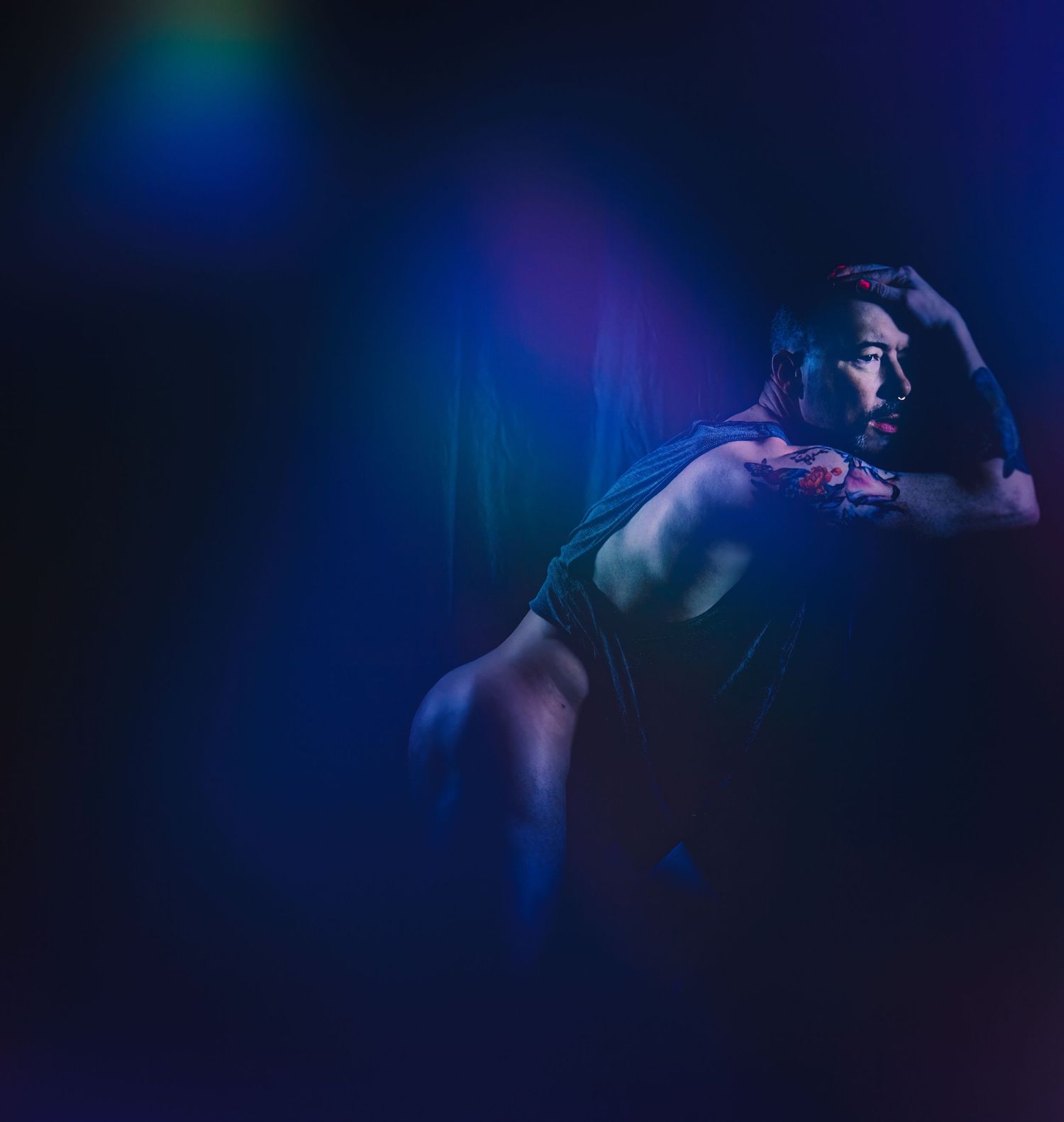 Dramatic blue and purple lit portrait of muscular man resting hand on head in dark setting.
