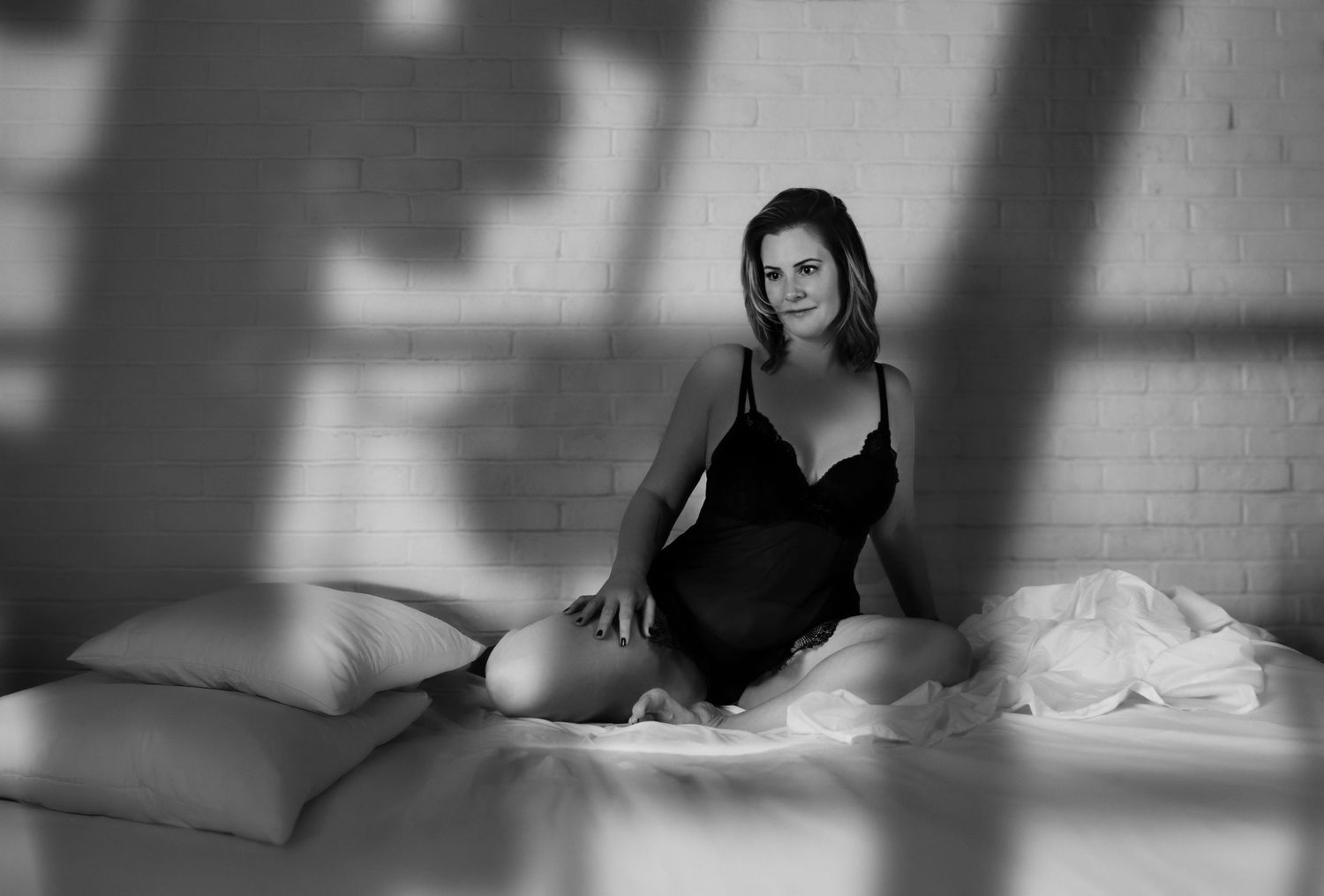 Black and white boudoir portrait of woman in black lingerie sitting on white bed with shadow patterns.