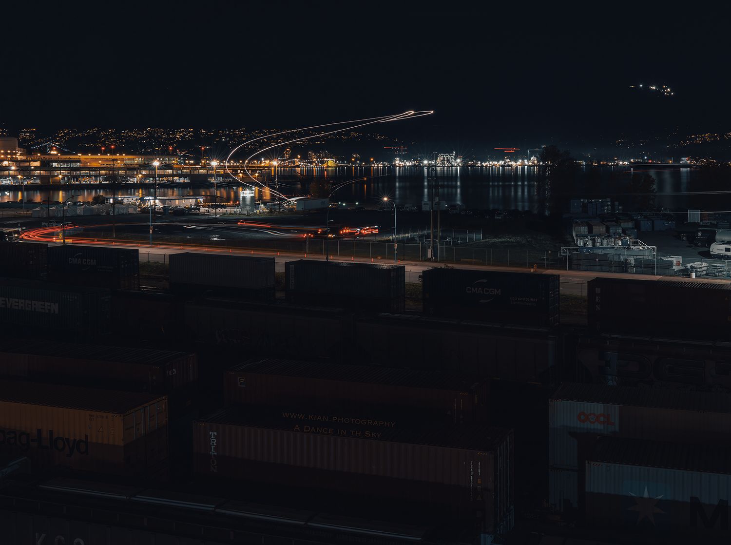 Aerial night view of a busy shipping container port with city lights reflecting on water in the background.