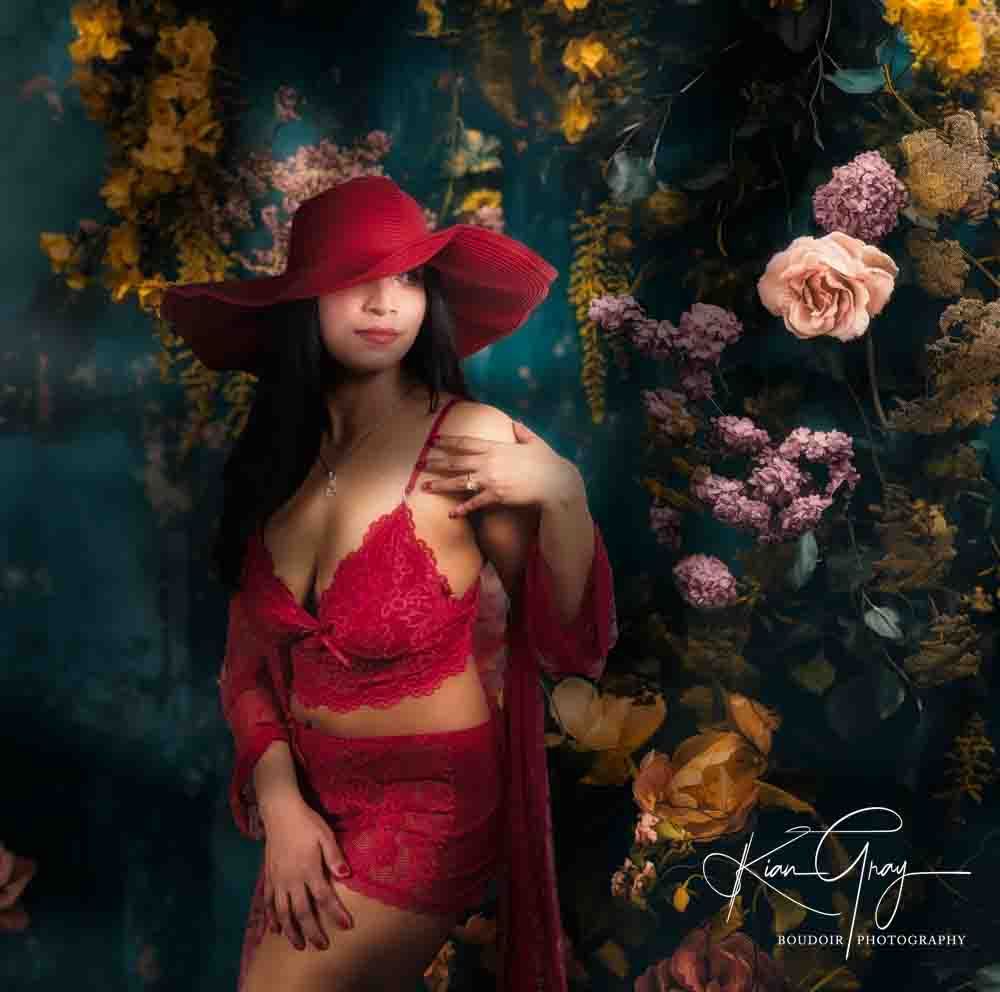 Woman in red lace lingerie and floppy red hat poses among colorful floral backdrop in boudoir portrait.