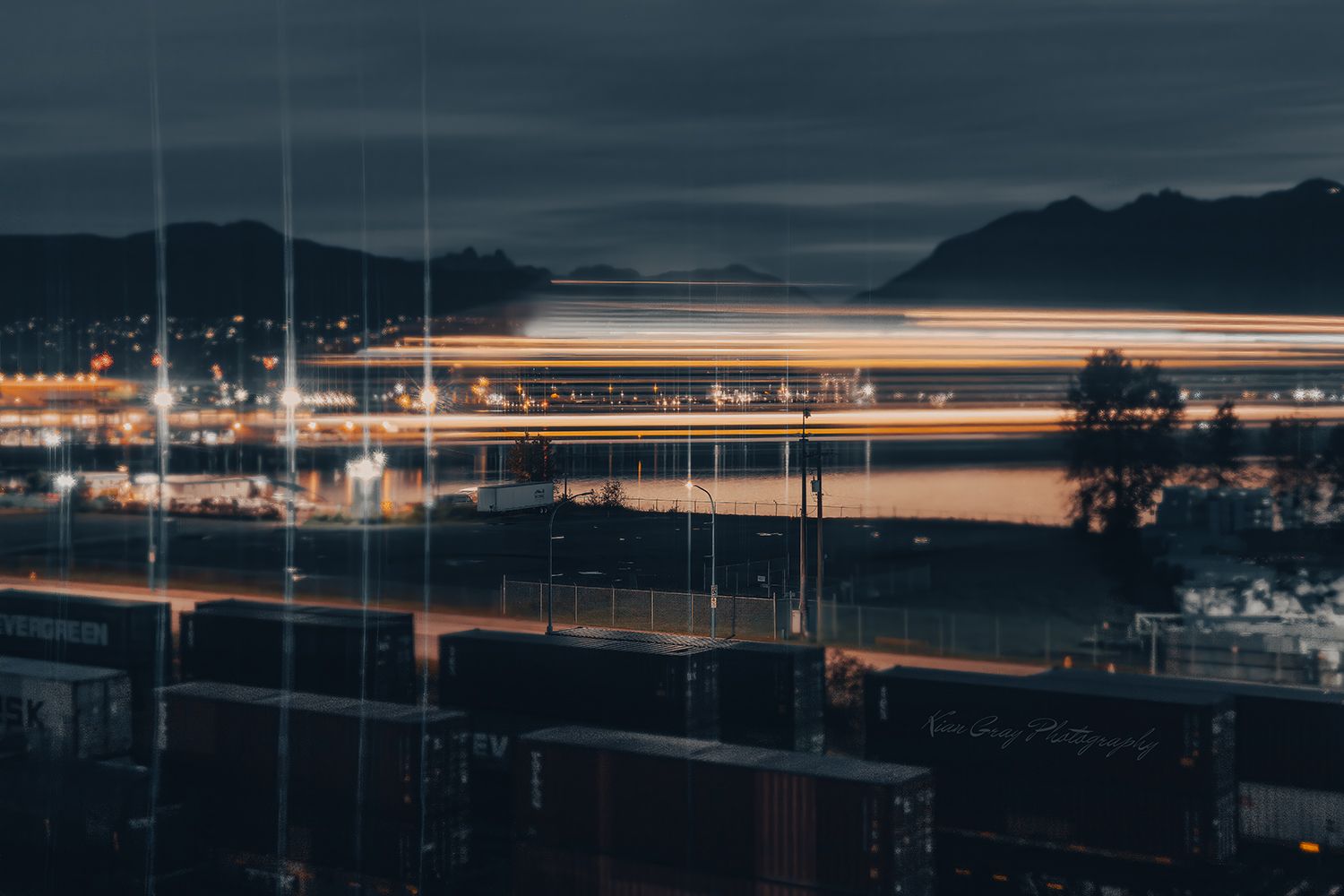 Long exposure night shot of a busy port with glowing light trails reflecting on water, mountains silhouetted in background.