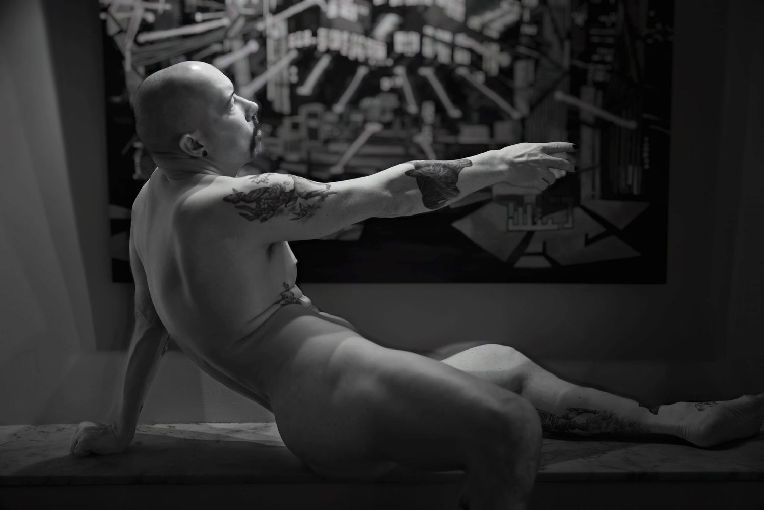 Black and white artistic nude photo of a tattooed bald man posing near a framed artwork.