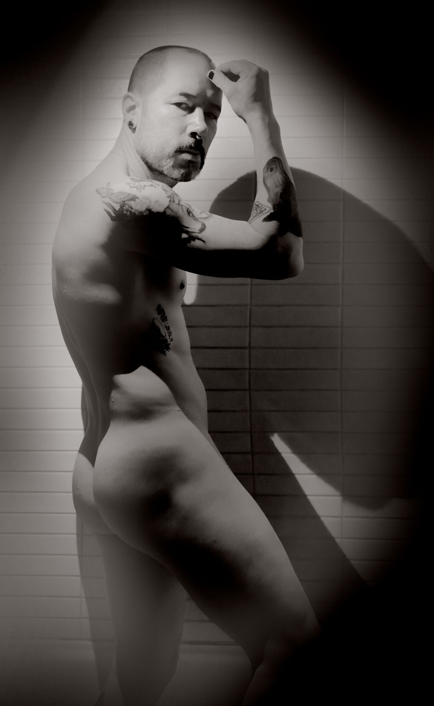 Artistic black and white nude portrait of a tattooed Asian man posing against a tiled wall.