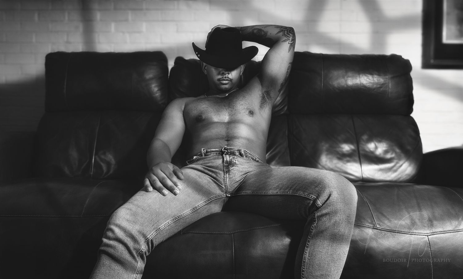 Black and white photo of shirtless man in jeans relaxing on leather sofa wearing cowboy hat.