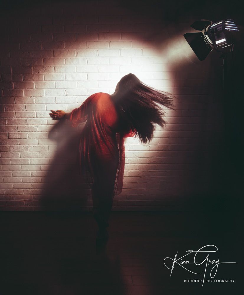 Dramatic boudoir photo of woman in red dress under spotlight against brick wall with hair in motion.