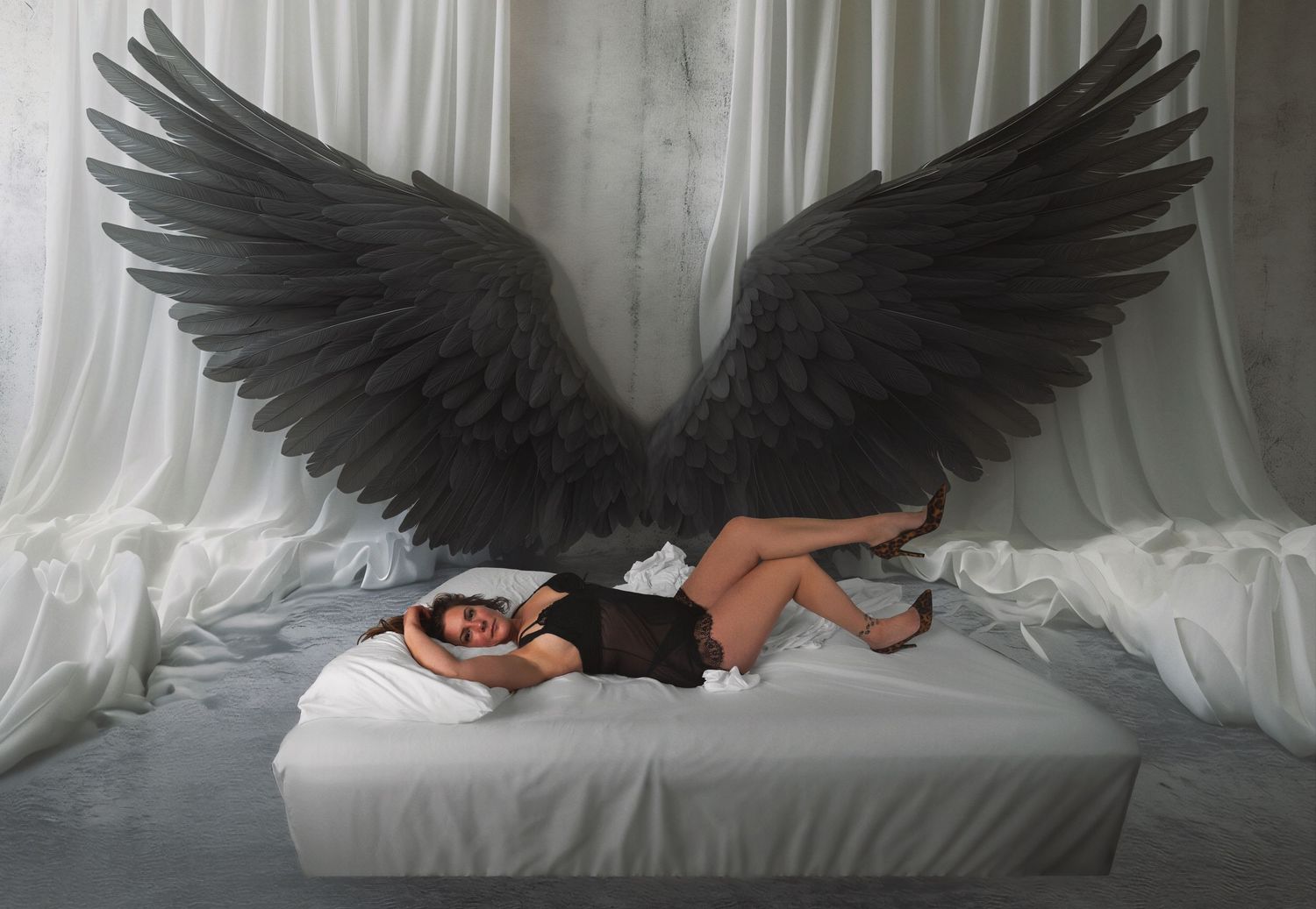 Woman in black dress lying on white bed with large dark angel wings backdrop.