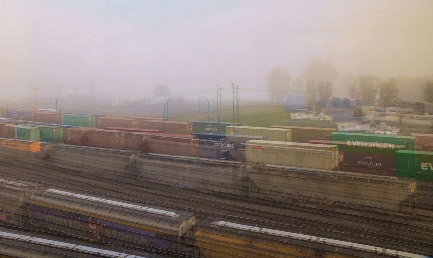 Freight trains with colorful shipping containers navigate a foggy rail yard on a misty morning.