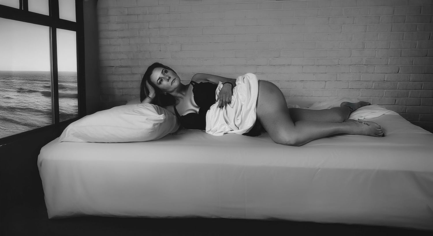 Black and white boudoir photo of woman lying on white bed with ocean view window in background.