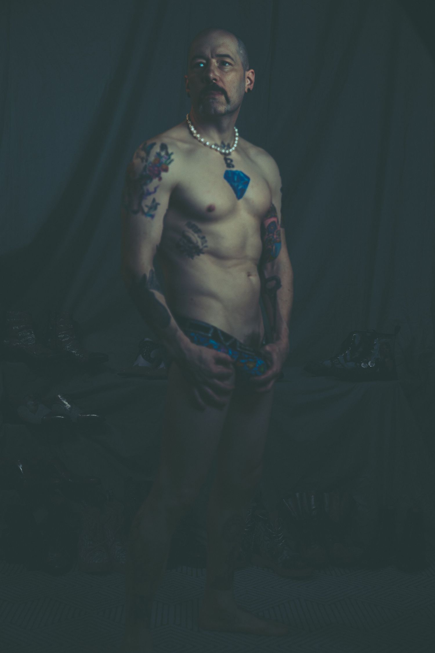 A tattooed shirtless bald man with a pearl necklace stands in dramatic dark studio lighting.
