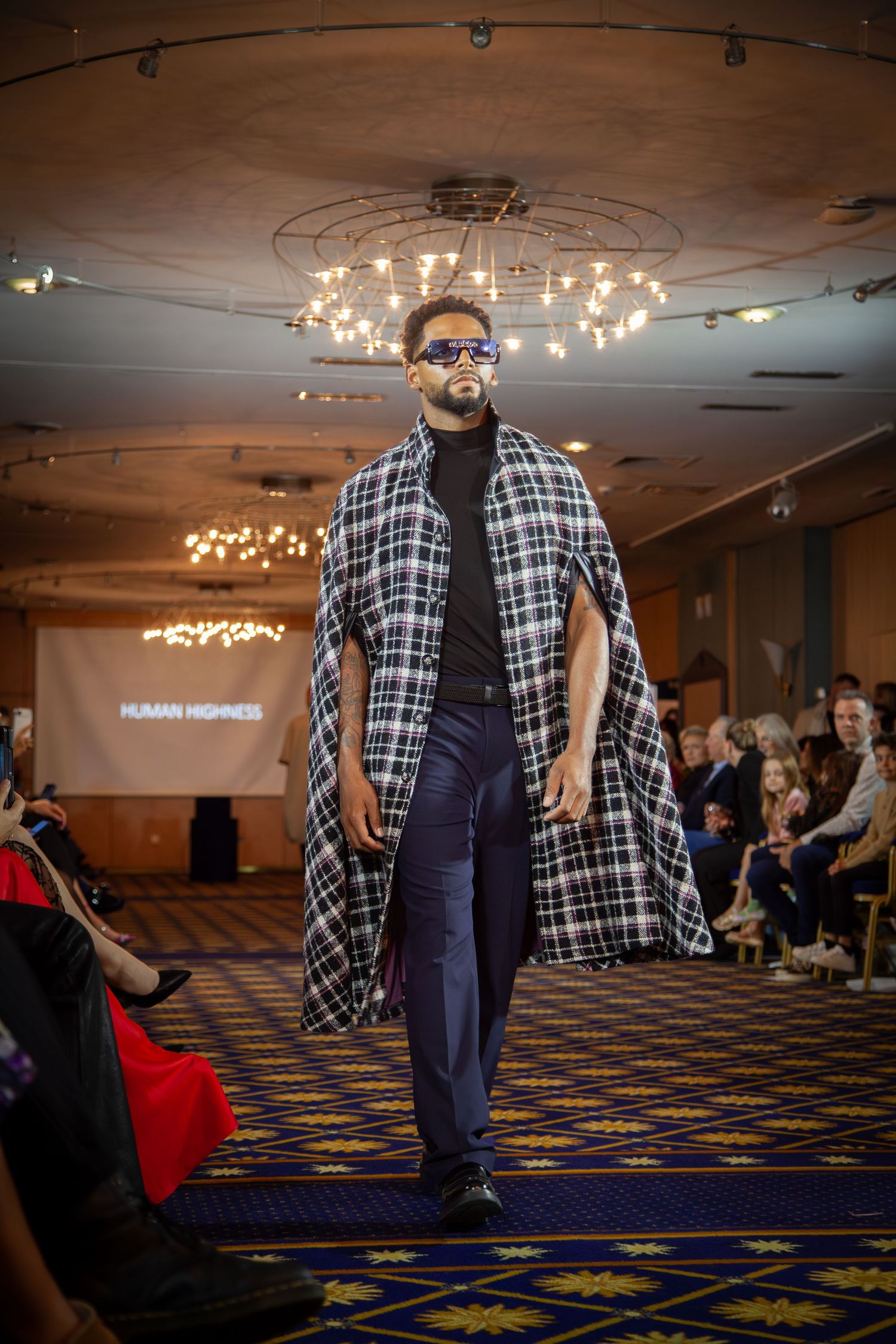 Male model walks runway in plaid cape, black turtleneck, navy pants, and blue sunglasses at Human Highness fashion show.