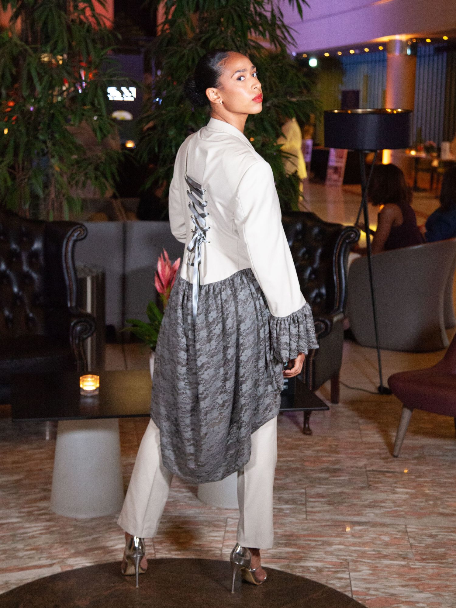 Model poses in white jacket with gray lace overlay skirt and white trousers at an elegant fashion event.