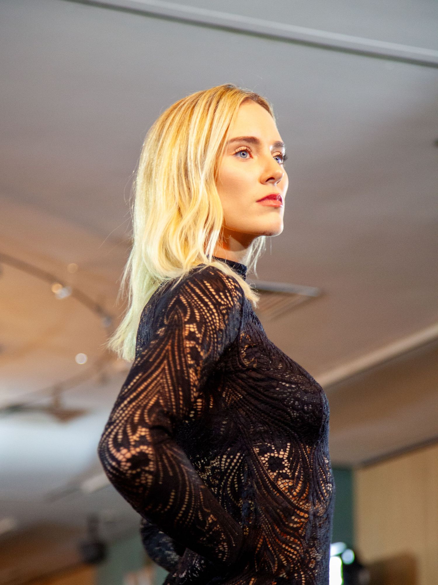 Blonde woman in black lace long-sleeve dress poses elegantly in a well-lit modern interior space.