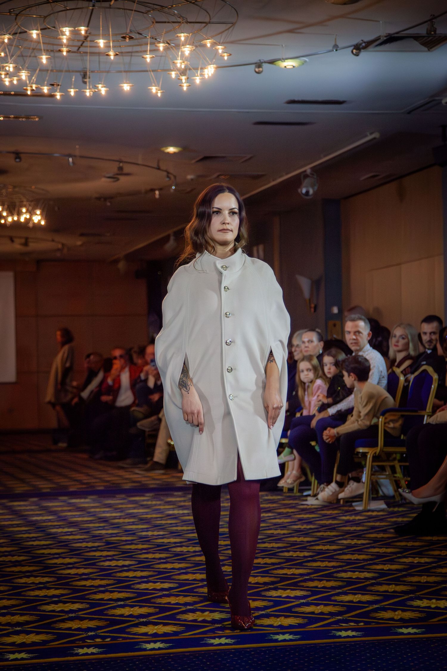 A model walks the runway in a white cape coat with burgundy tights at an indoor fashion show.