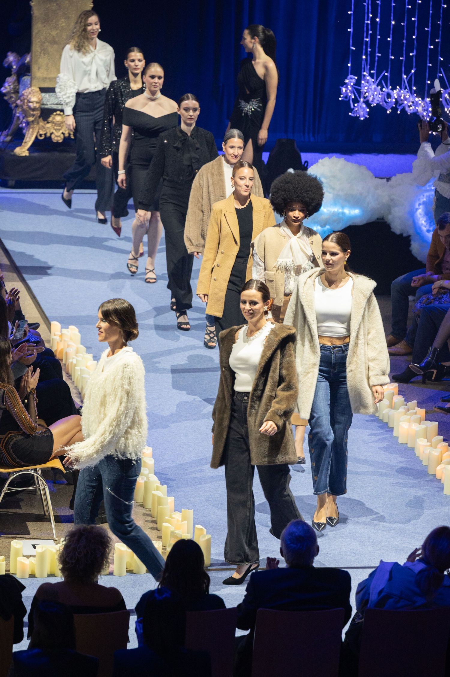 Models walk the runway at a fashion show, wearing elegant outfits, with candles lining the catwalk and audience watching.