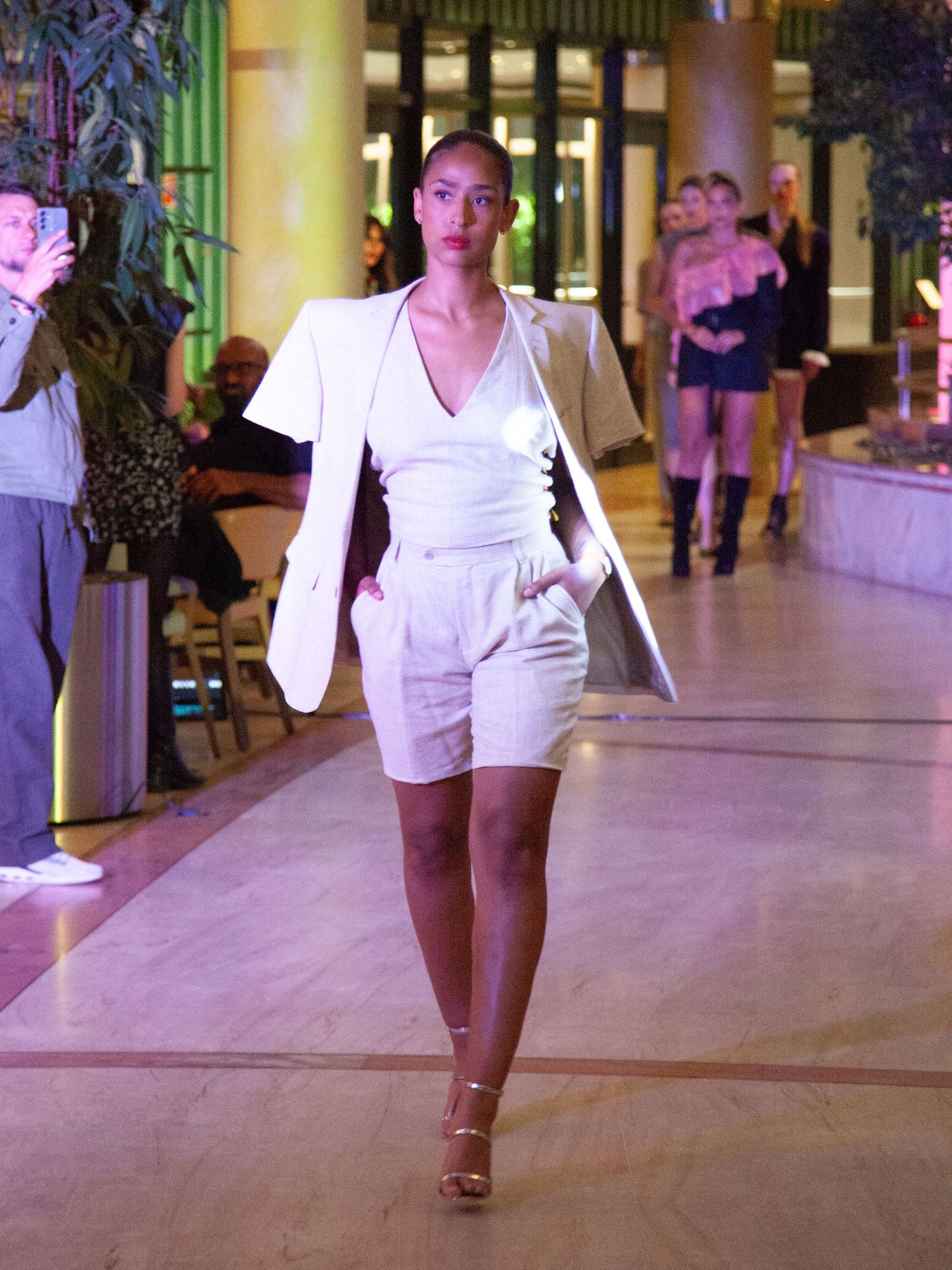 Model walks runway in white blazer, shorts, and heels at an indoor fashion show.