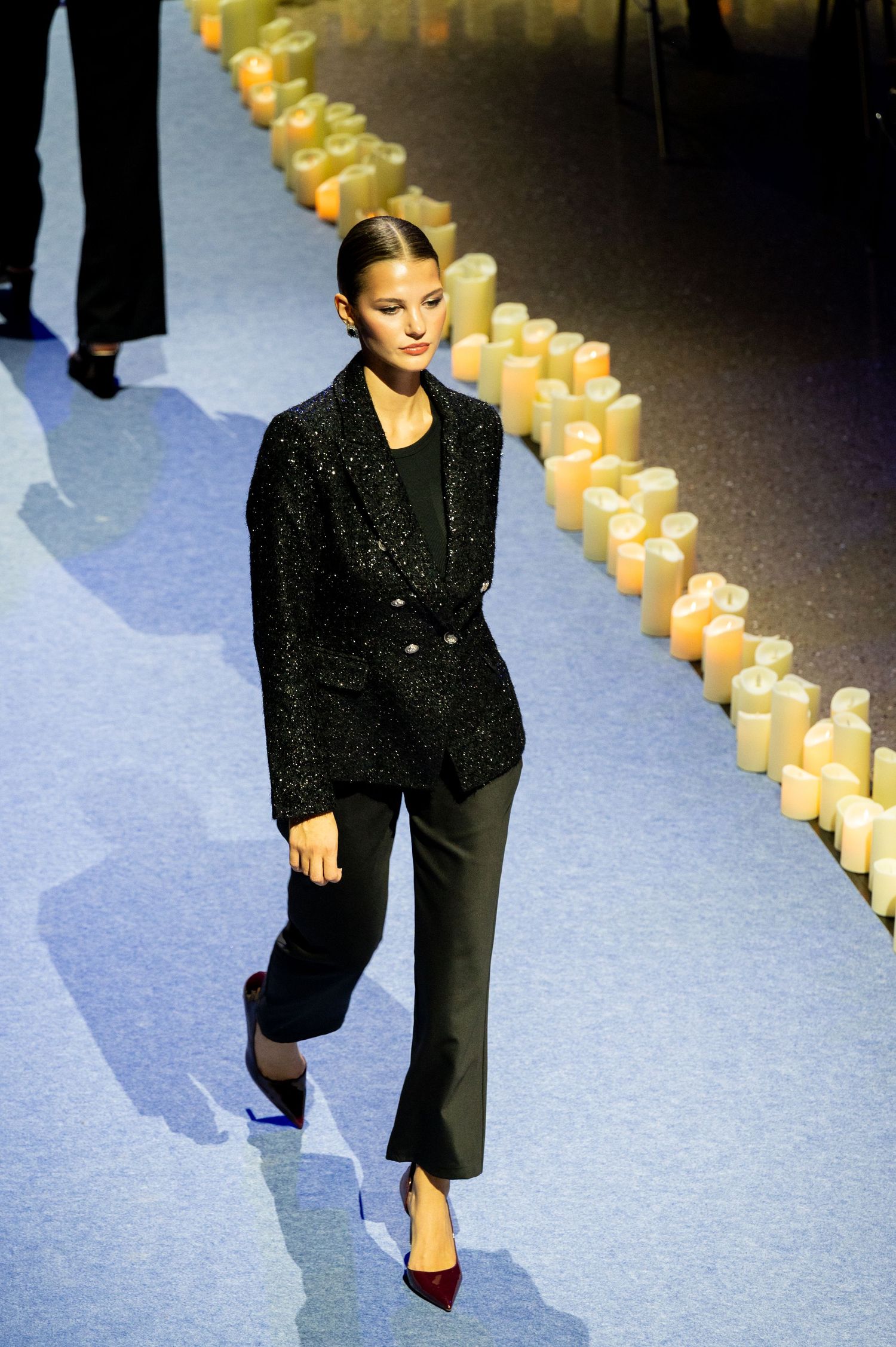 Model walks runway in black sequin blazer and trousers with red heels, flanked by glowing candles.