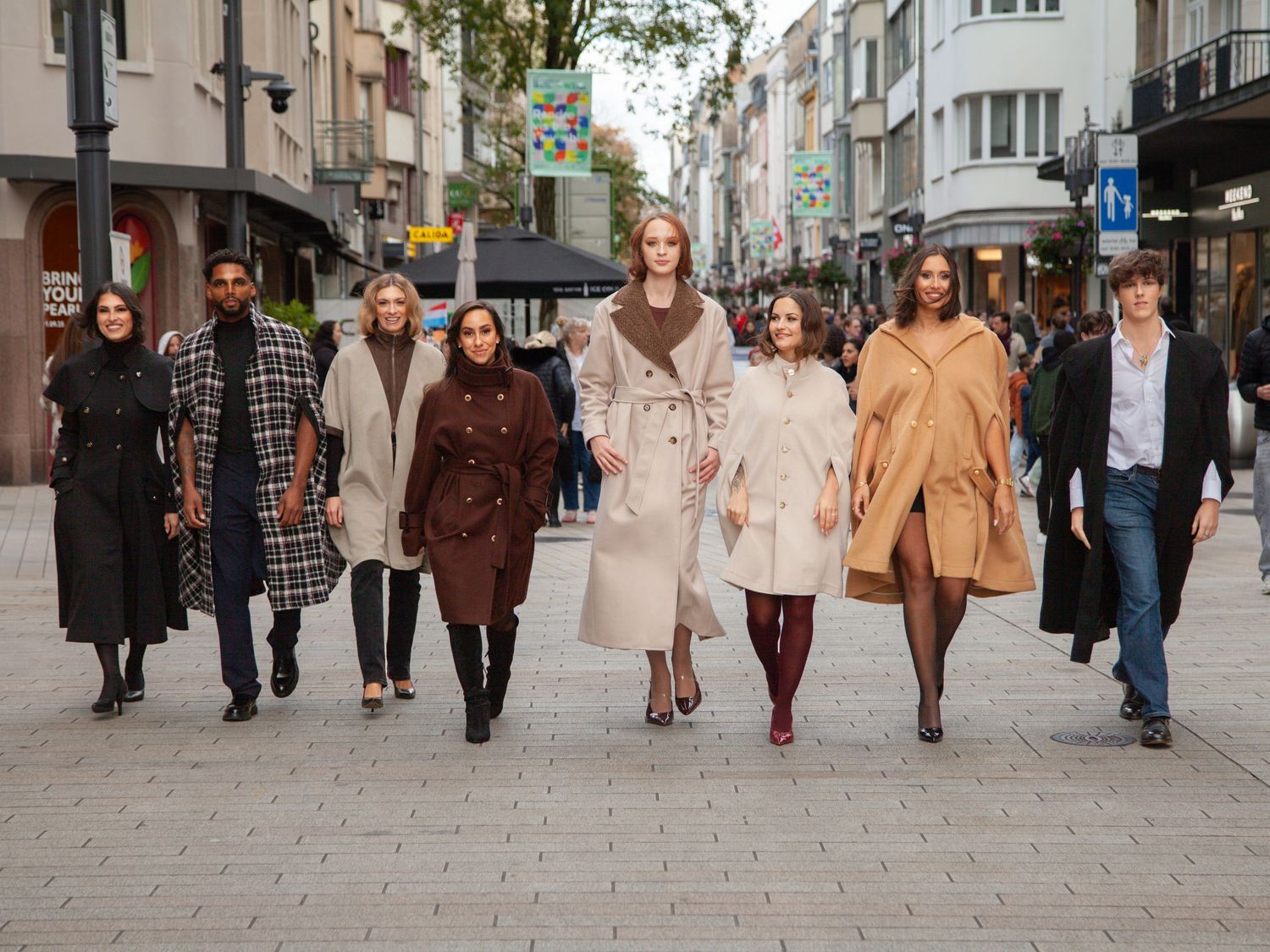Group of diverse models walking confidently in stylish autumn coats on a busy European shopping street.