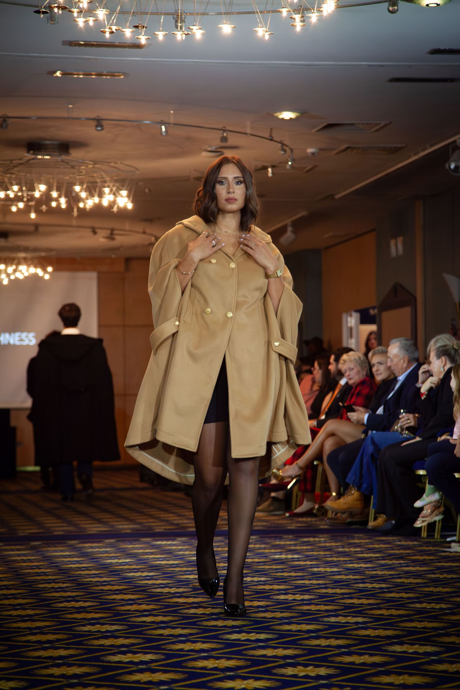 Model walks runway in camel cape coat at fashion show, with seated audience watching in elegant venue.