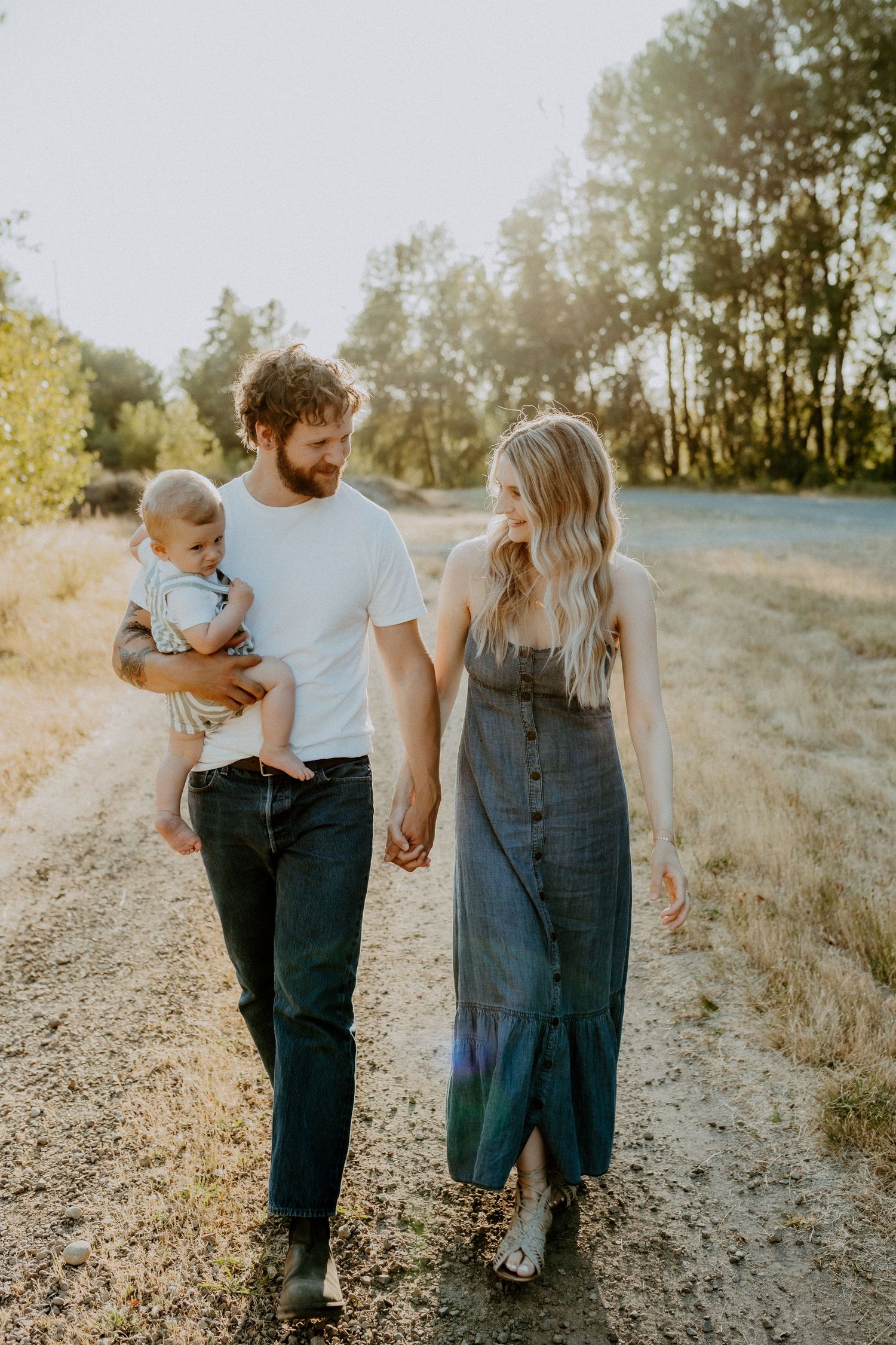 A young family walks together on a sunlit dirt path surrounded by trees.