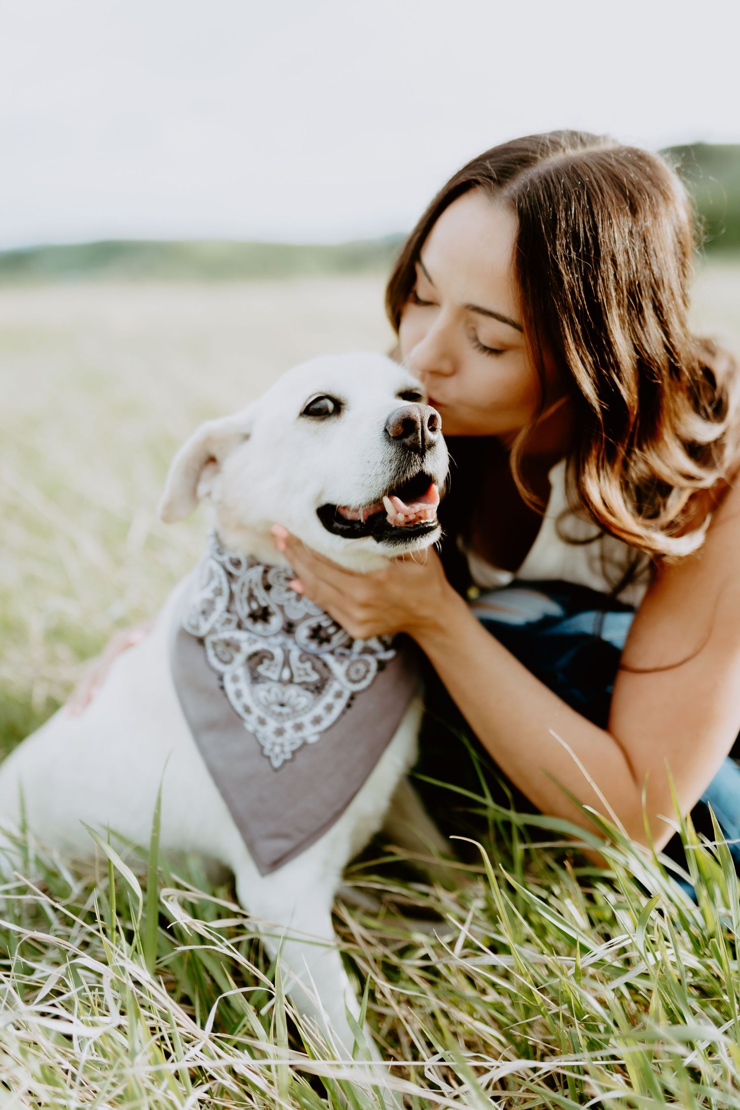 Woman cuddling with white dog wearing bandana in sunny meadow.