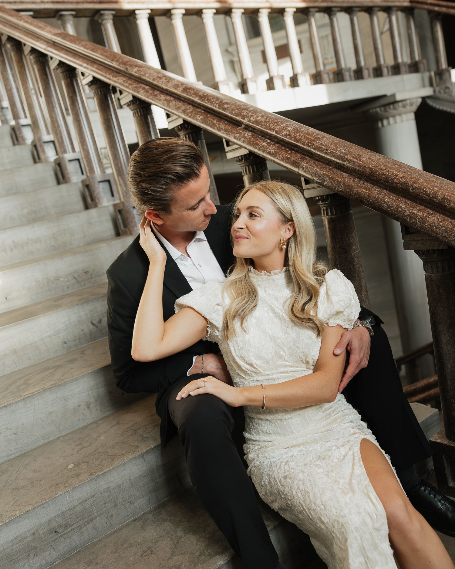 A couple in formal attire share an intimate moment while sitting on wooden stairs in a vintage setting.