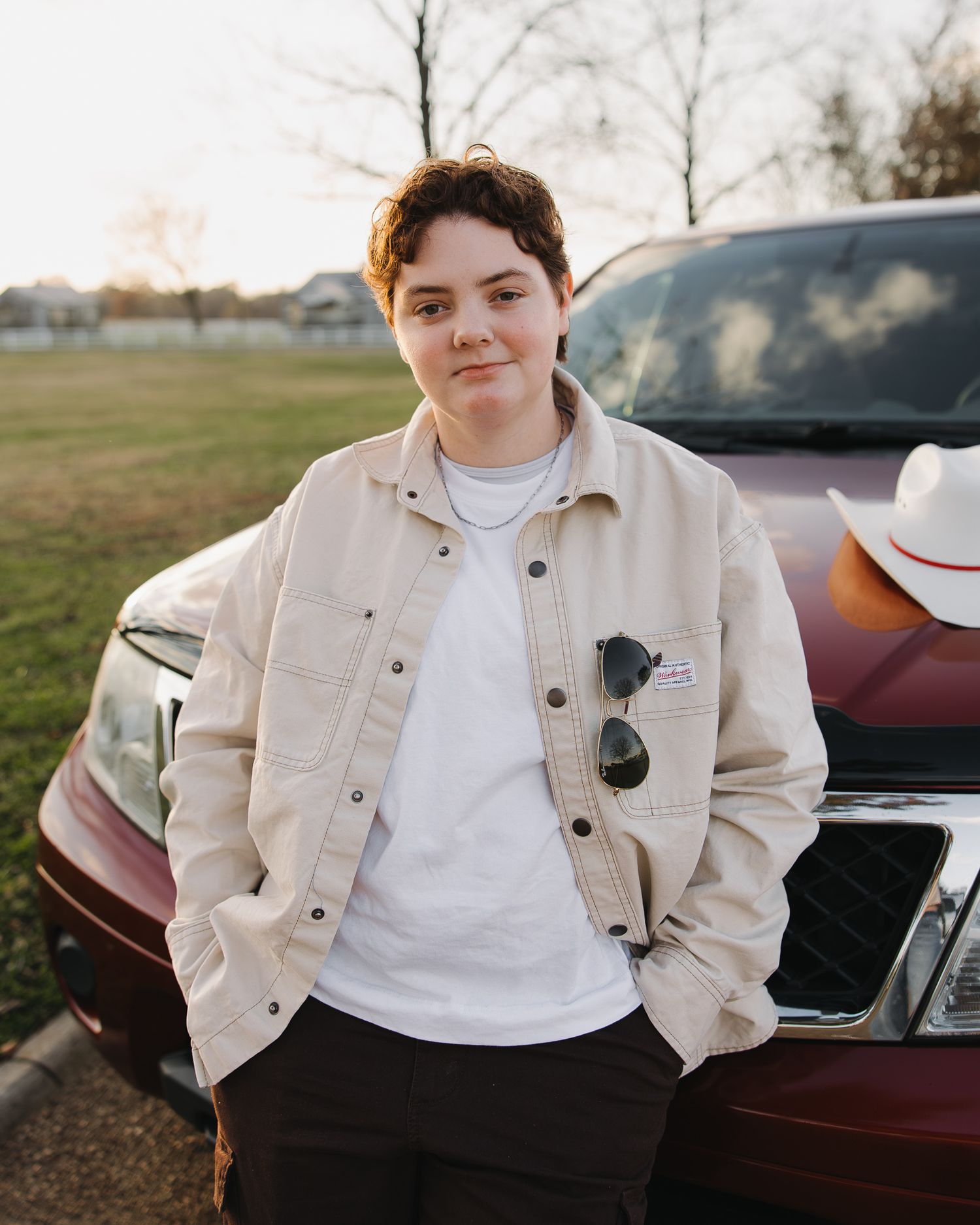 A person in a beige denim jacket and white t-shirt poses casually against a car in a rural outdoor setting at sunset photography