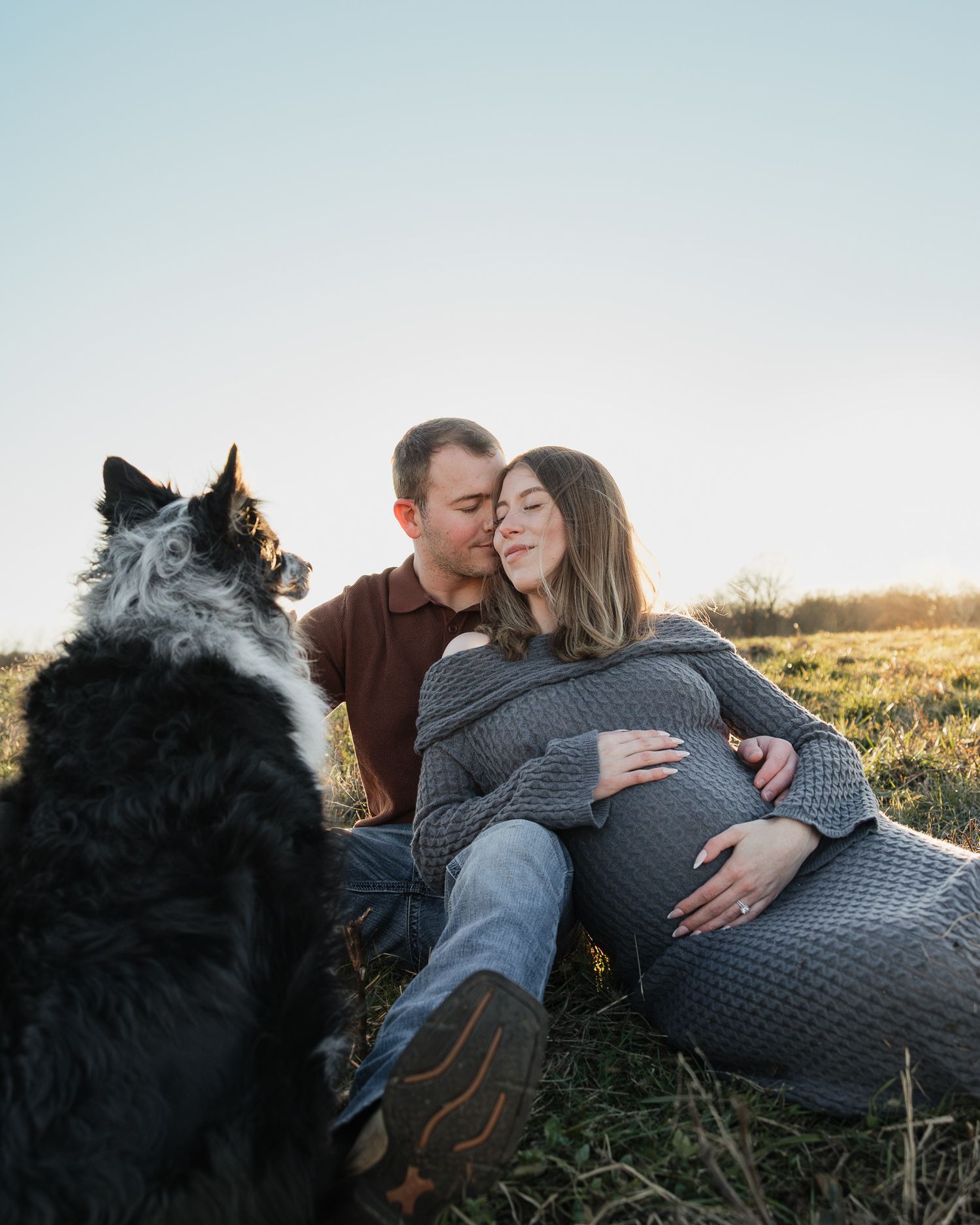 Intimate maternity session in a field at golden hour with a black dog watching nearby.