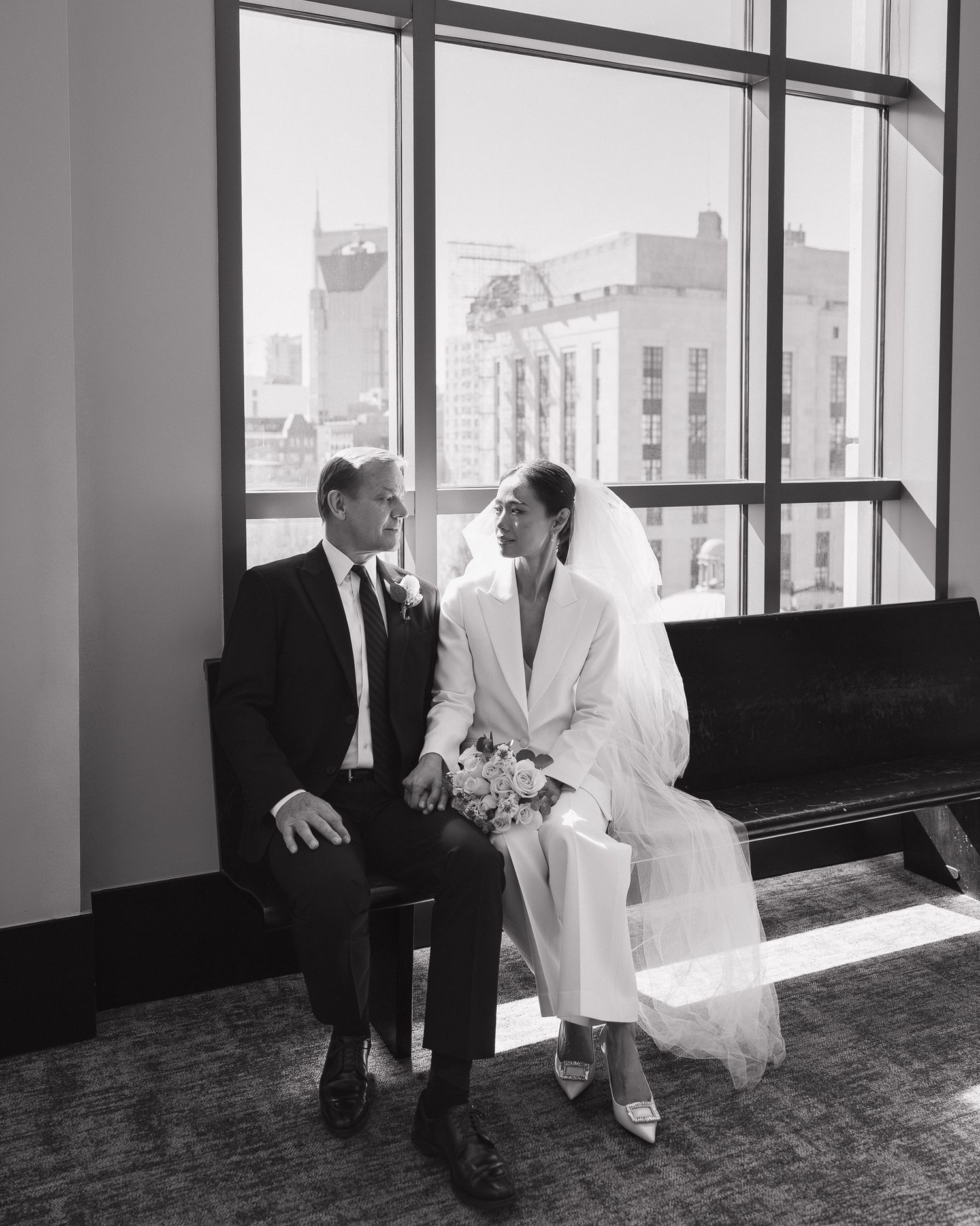 Black and white wedding photo of bride in white suit and groom in tuxedo sitting together by large windows.