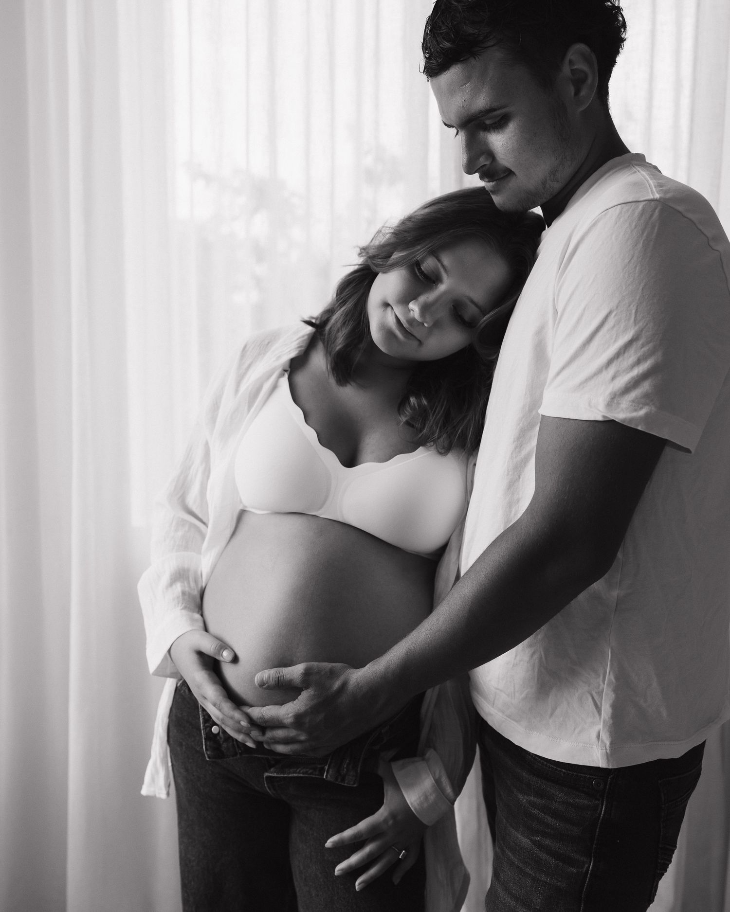 Black and white maternity photo of a couple tenderly cradling a pregnant belly near a bright window.
