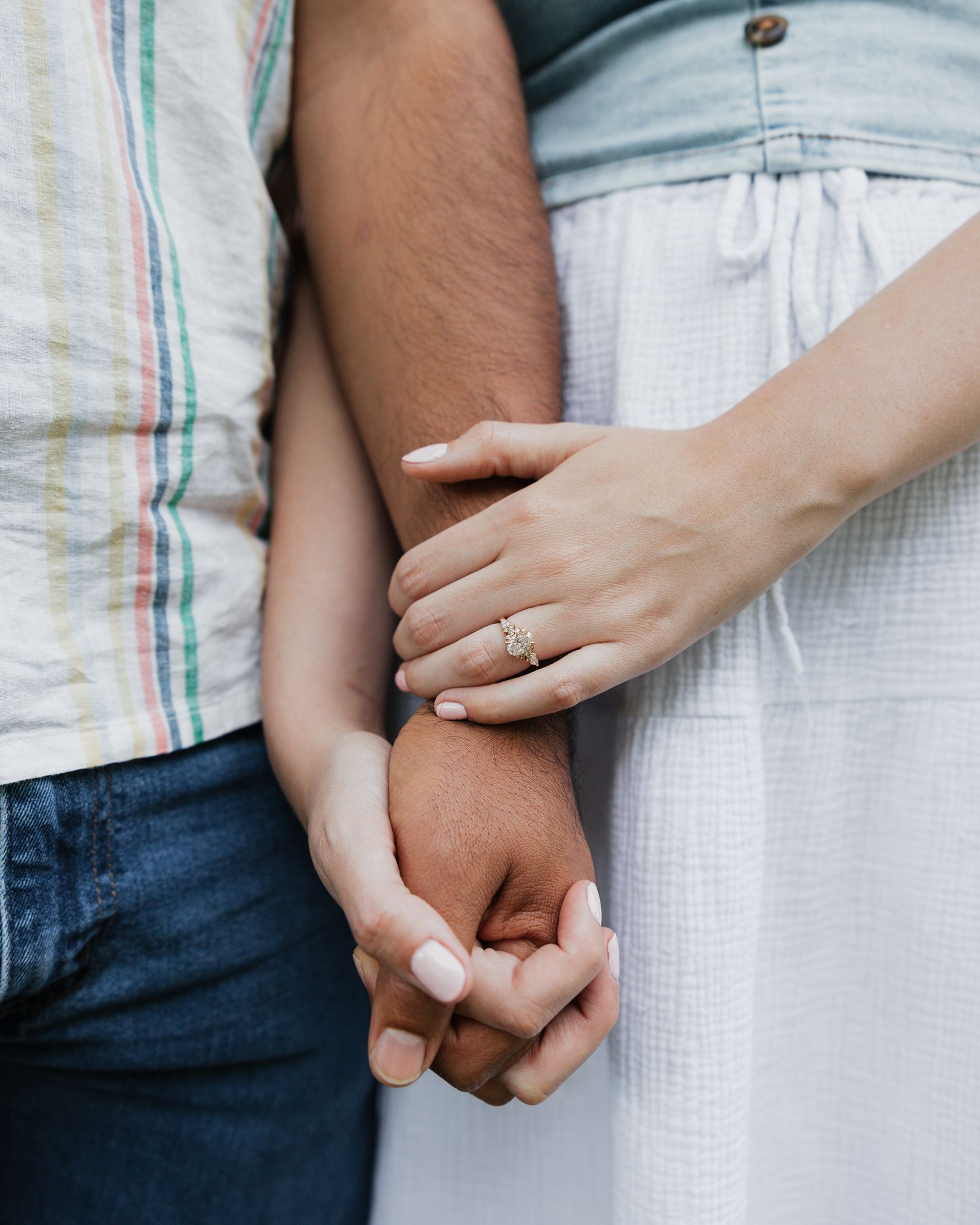 A couple holds hands tenderly, the woman wearing an engagement ring, dressed in casual summer attire.