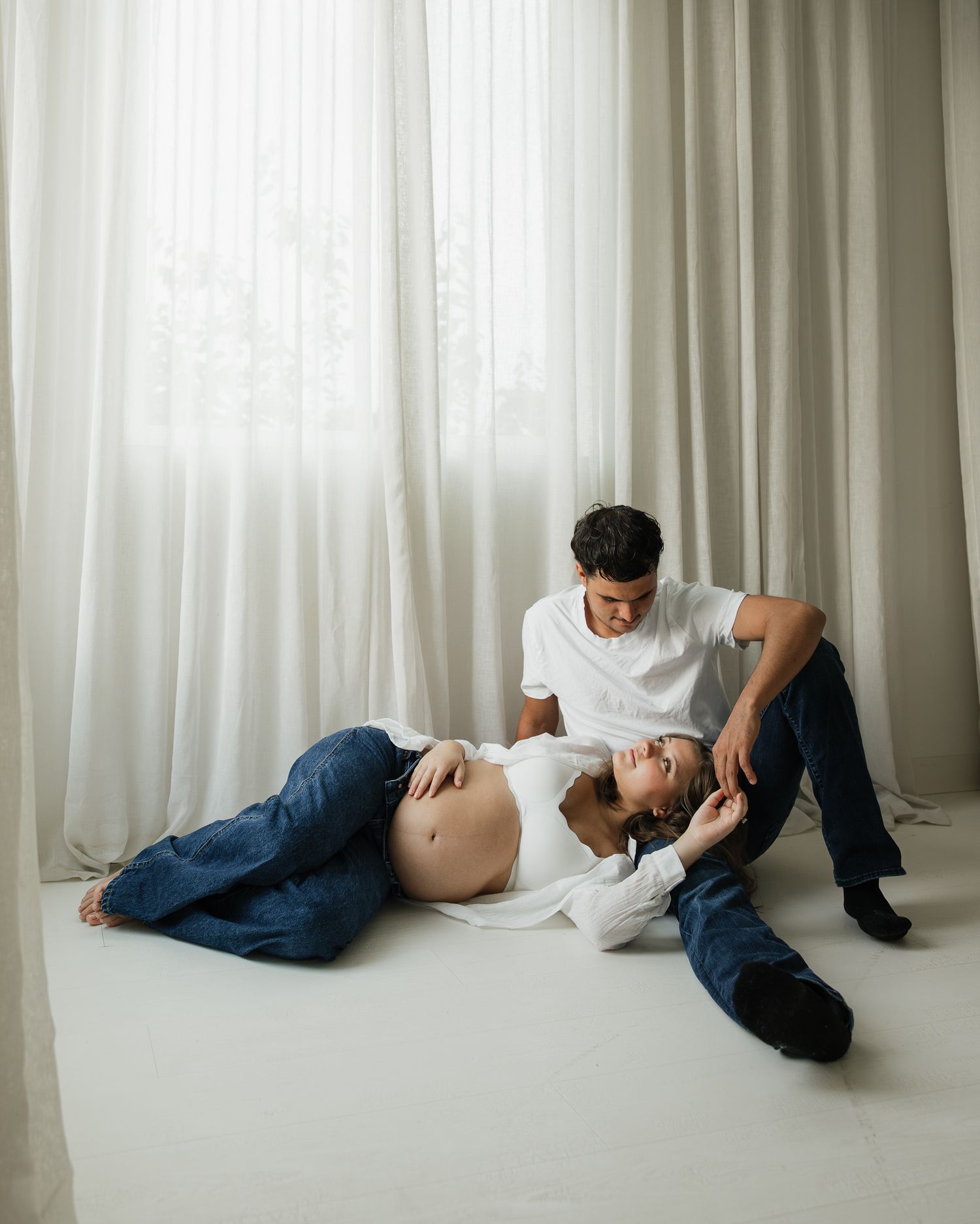 Couple poses for maternity photo shoot, pregnant woman lying on floor with partner sitting beside her.