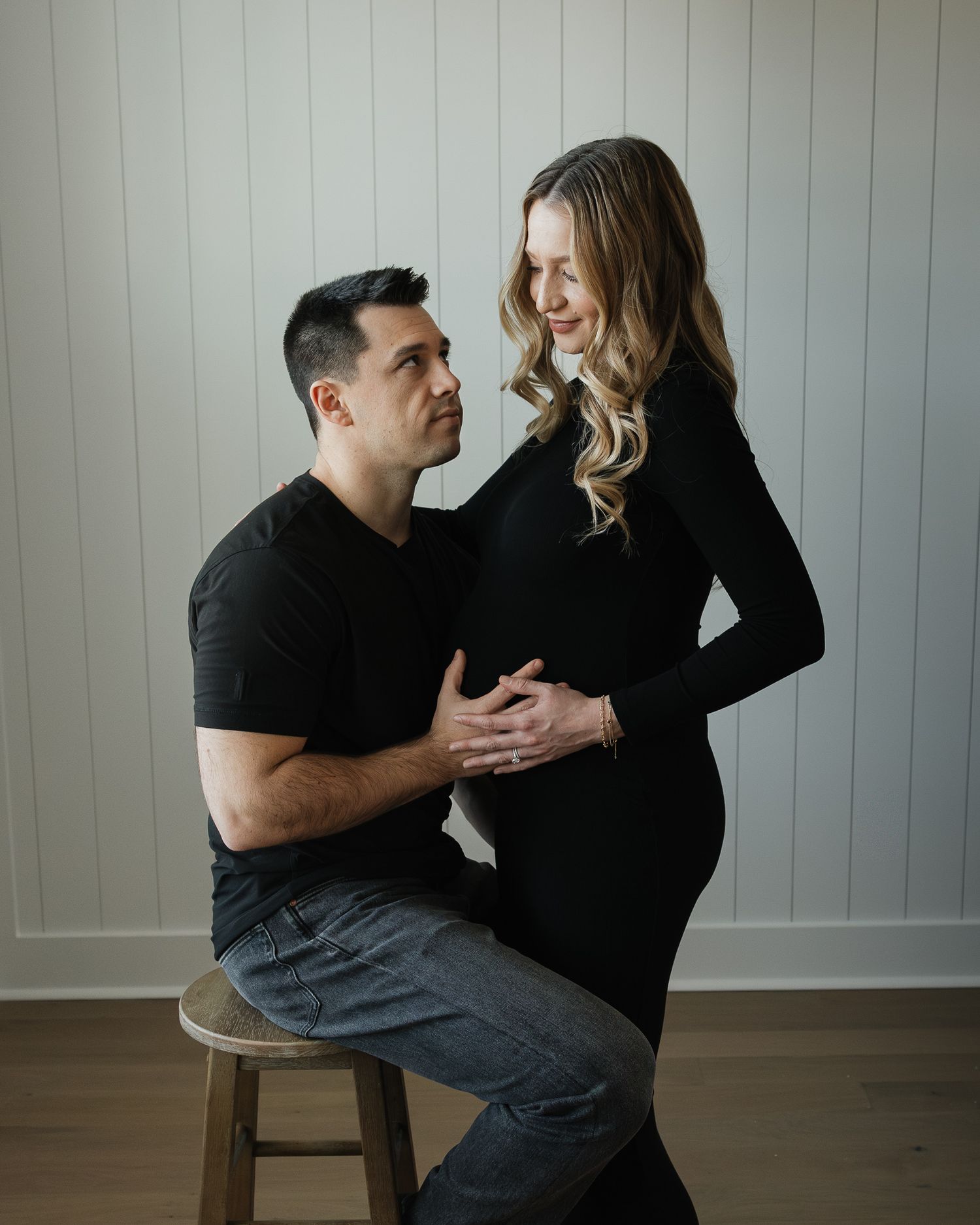 A couple shares a candid moment together while seated on a wooden chair against a white wall.