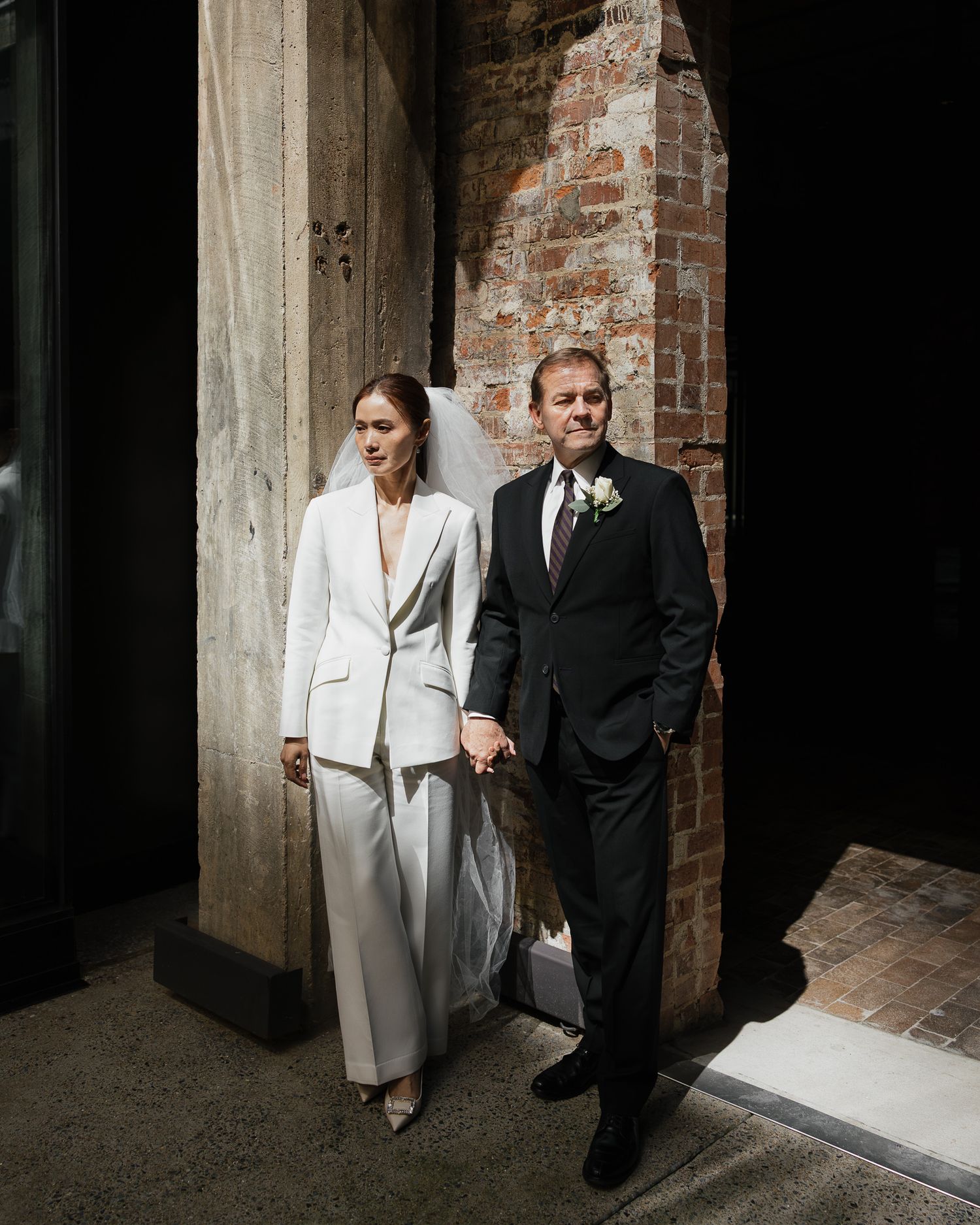 Bride in white pantsuit and groom in black suit holding hands outside rustic brick venue on wedding day in Downtown Nashville Tennessee.