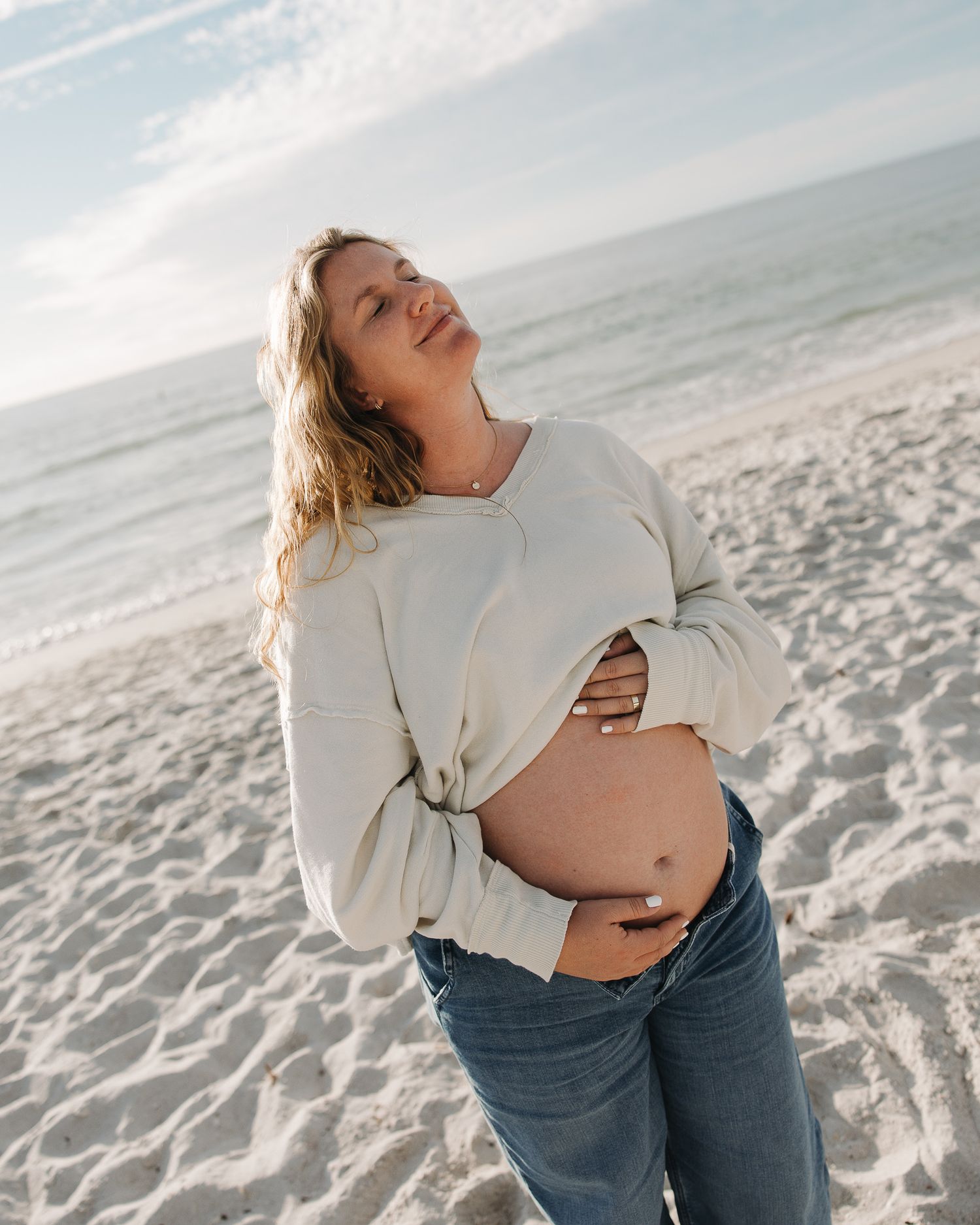 Maternity photos on sandy beach with person in cream sweater showing baby bump.