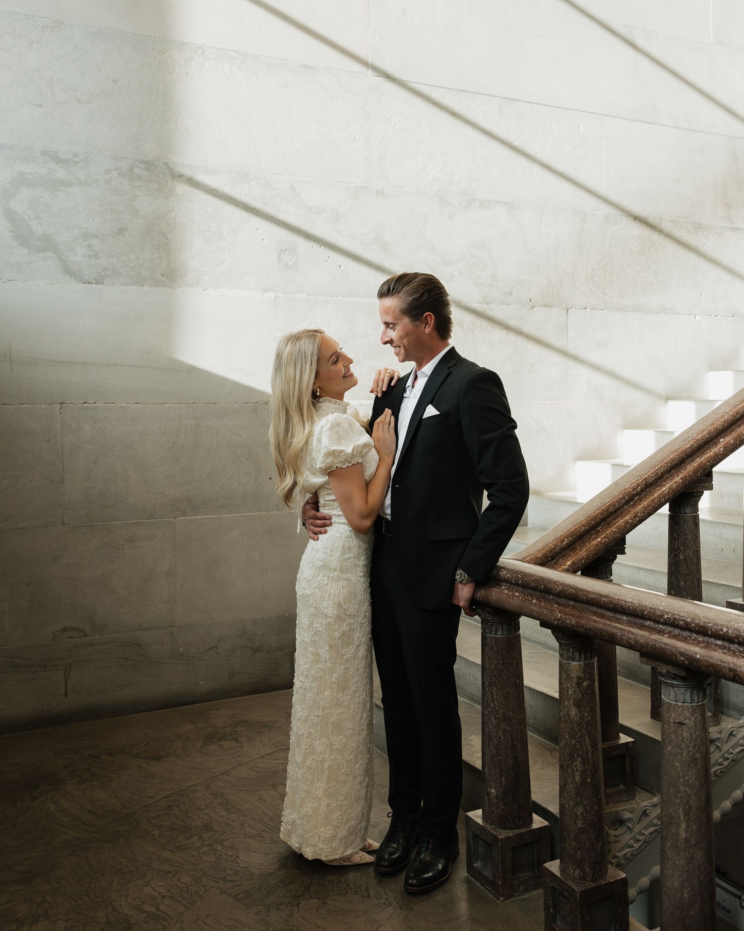A couple in nice attire share an intimate moment on a staircase with dramatic lighting and concrete walls for engagement photos