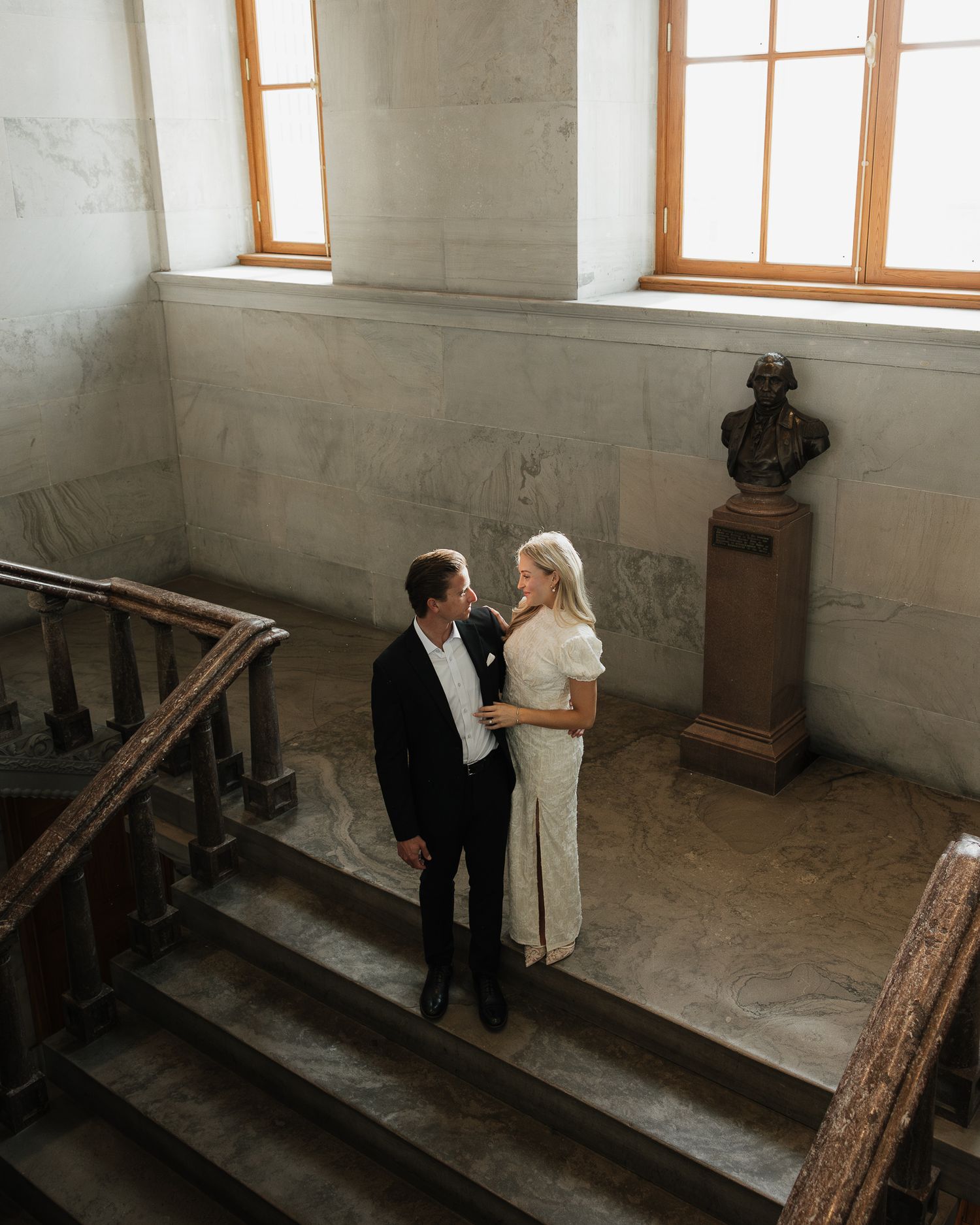 A couple stands intimately on a stone staircase in an old building with wooden windows and decorative pedestal.