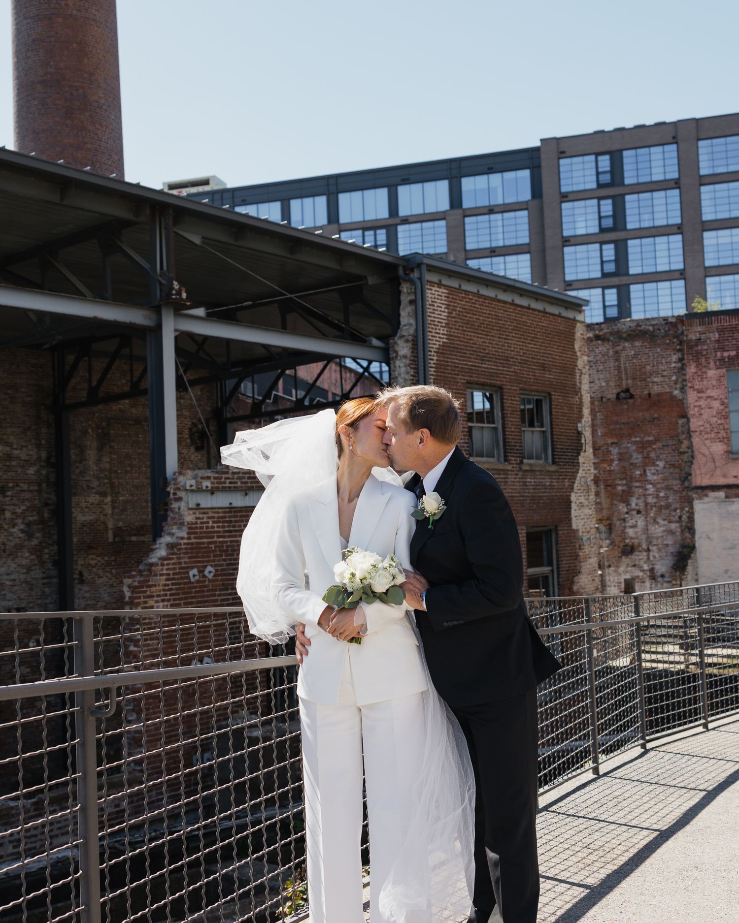 A bride in a white pantsuit and veil kisses her groom in a black tuxedo on a bridge at an urban venue.