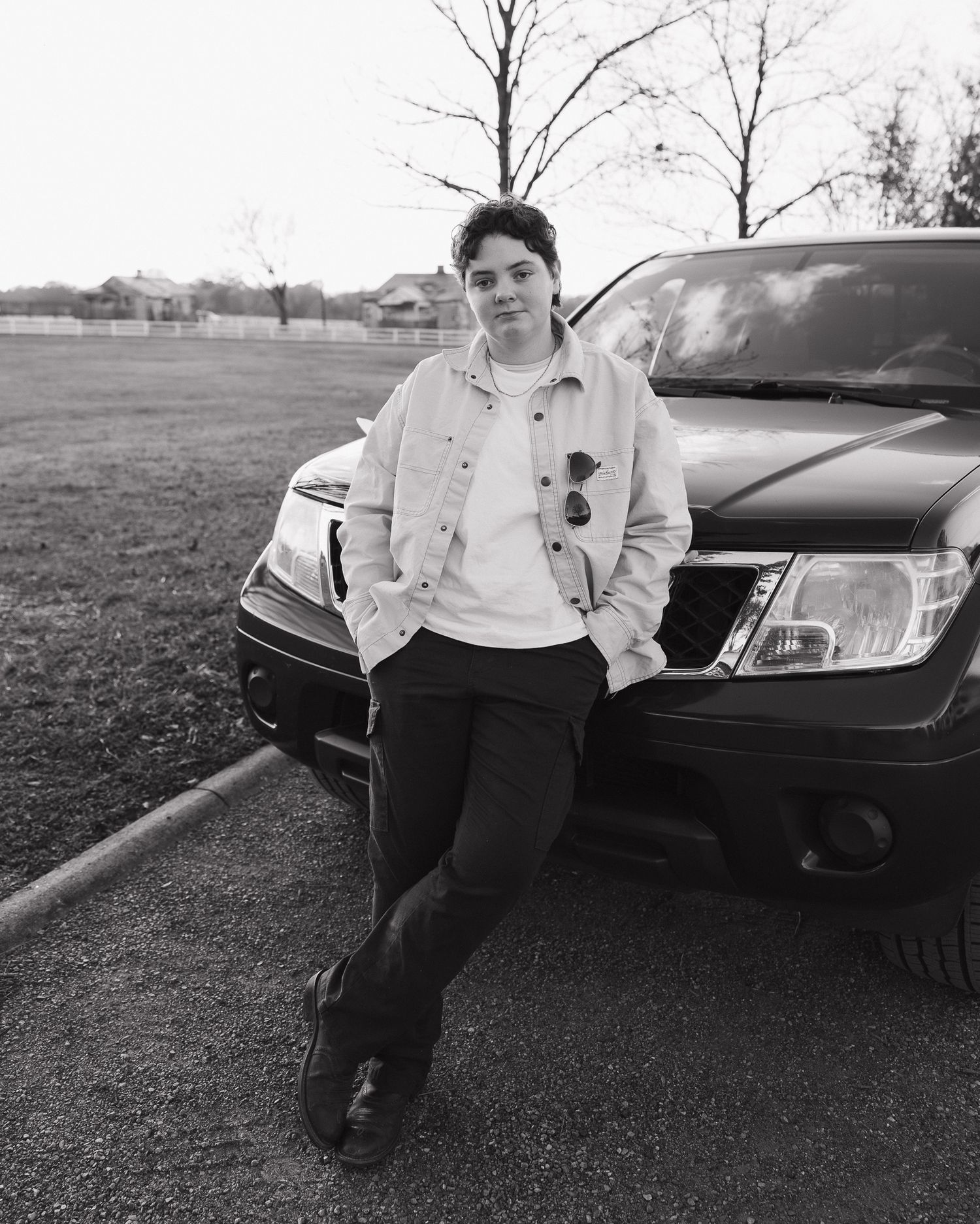 A person in a light colored sweatshirt sits casually against a dark car in an outdoor setting.