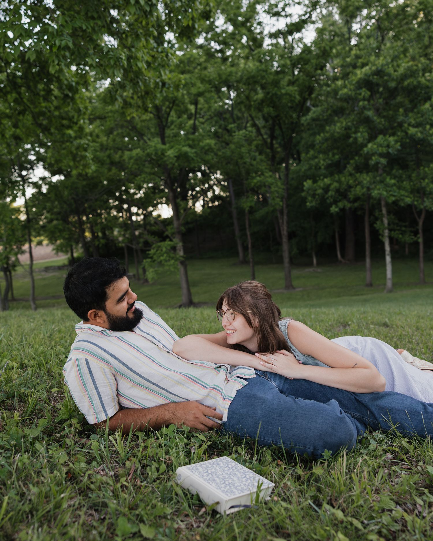 Couple relaxing on grass in a park, smiling at each other with a book nearby, surrounded by green trees.