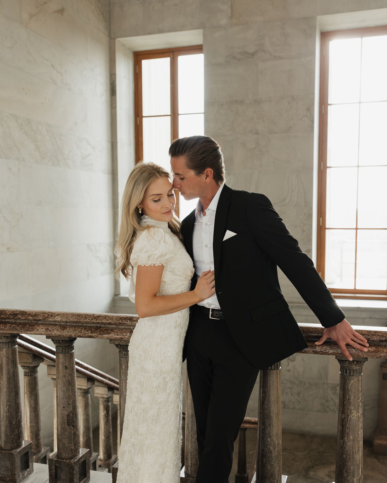 A couple in engagement attire share an intimate moment on an elegant staircase with natural light streaming through windows.
