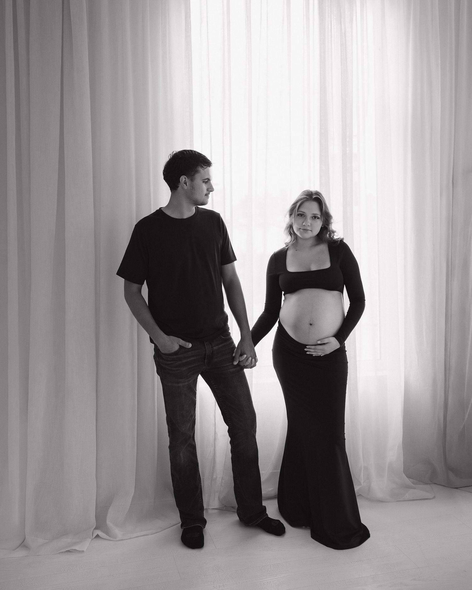 Black and white maternity photo of a couple holding hands, woman in black crop top and skirt cradling baby bump.