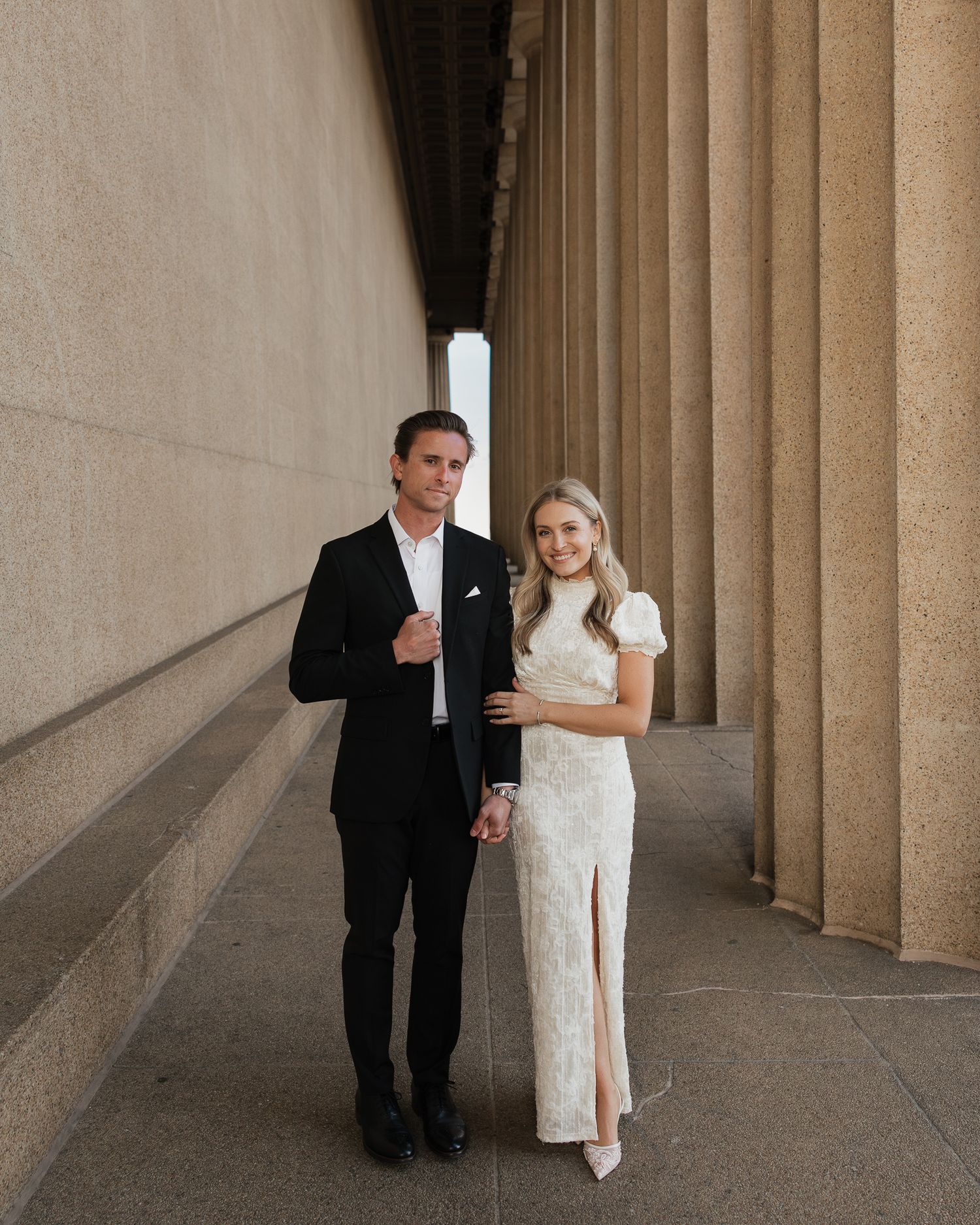 A couple in formal attire walks through an elegant colonnade with stone pillars casting dramatic shadows.