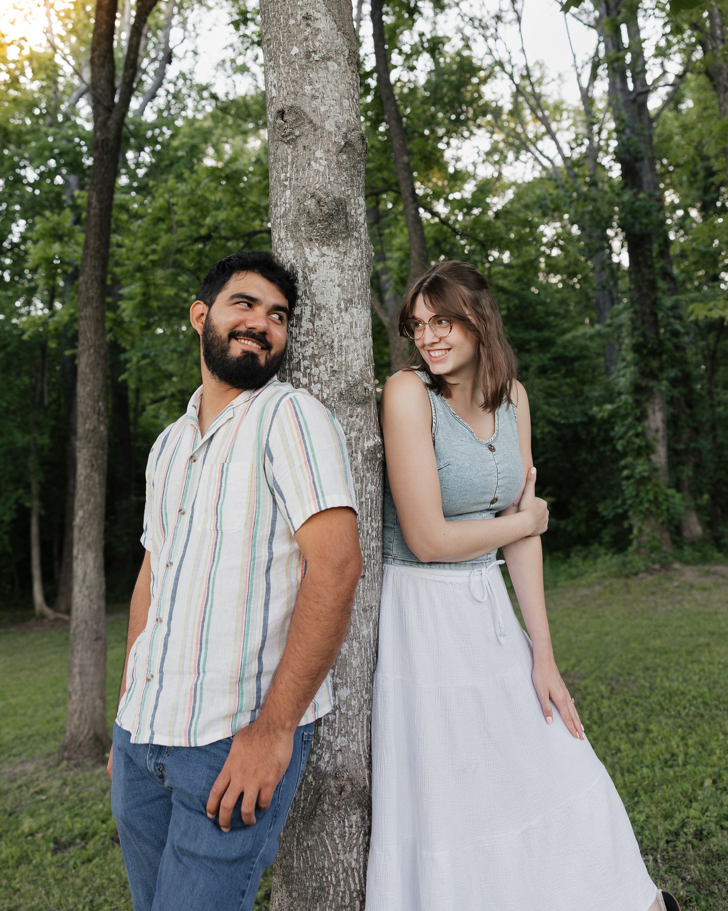 A bearded man and smiling woman lean back-to-back against a tree in a lush green park setting.