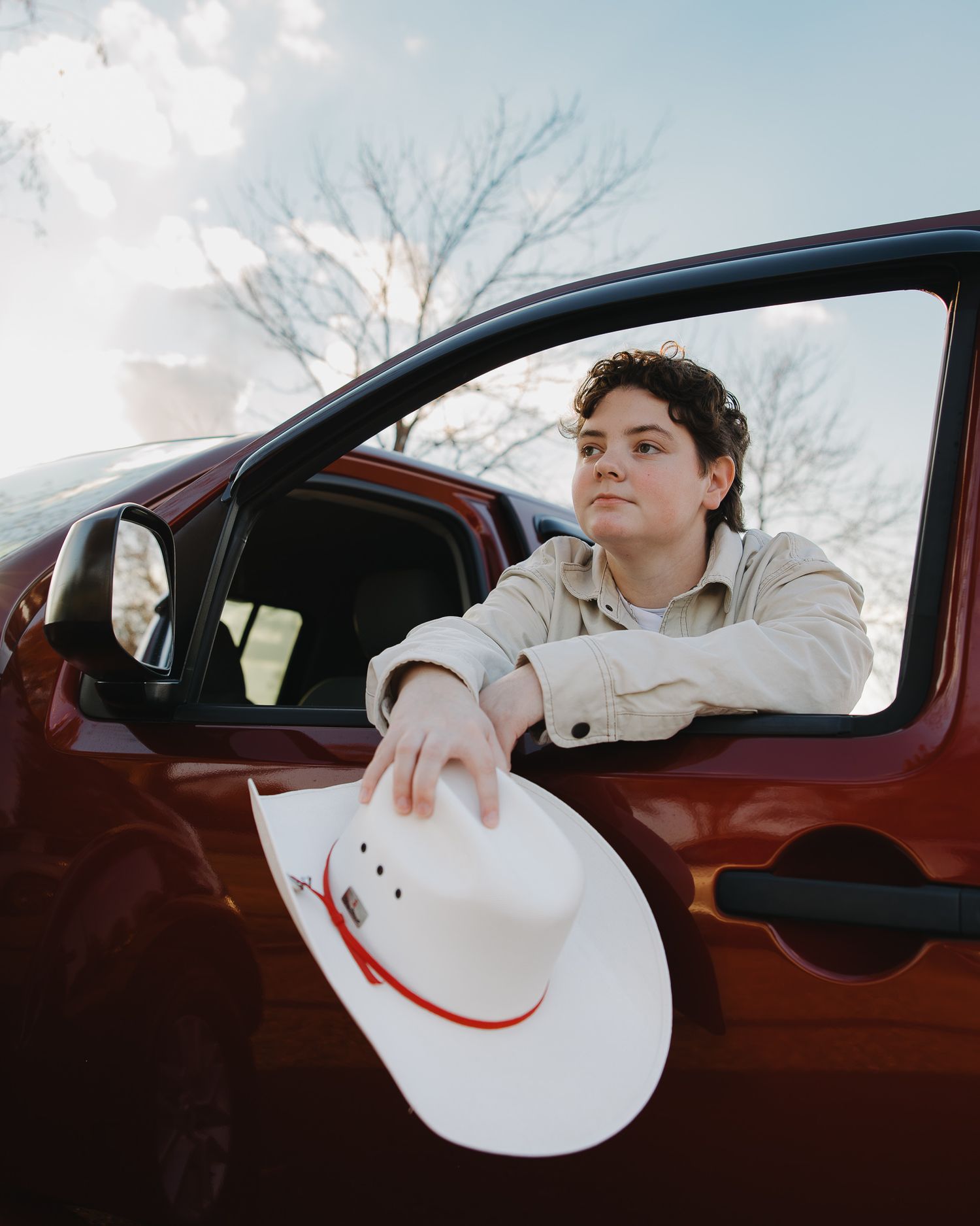 Someone sits in a red car holding a white cowboy hat with a smiley face design out the driver's side window.