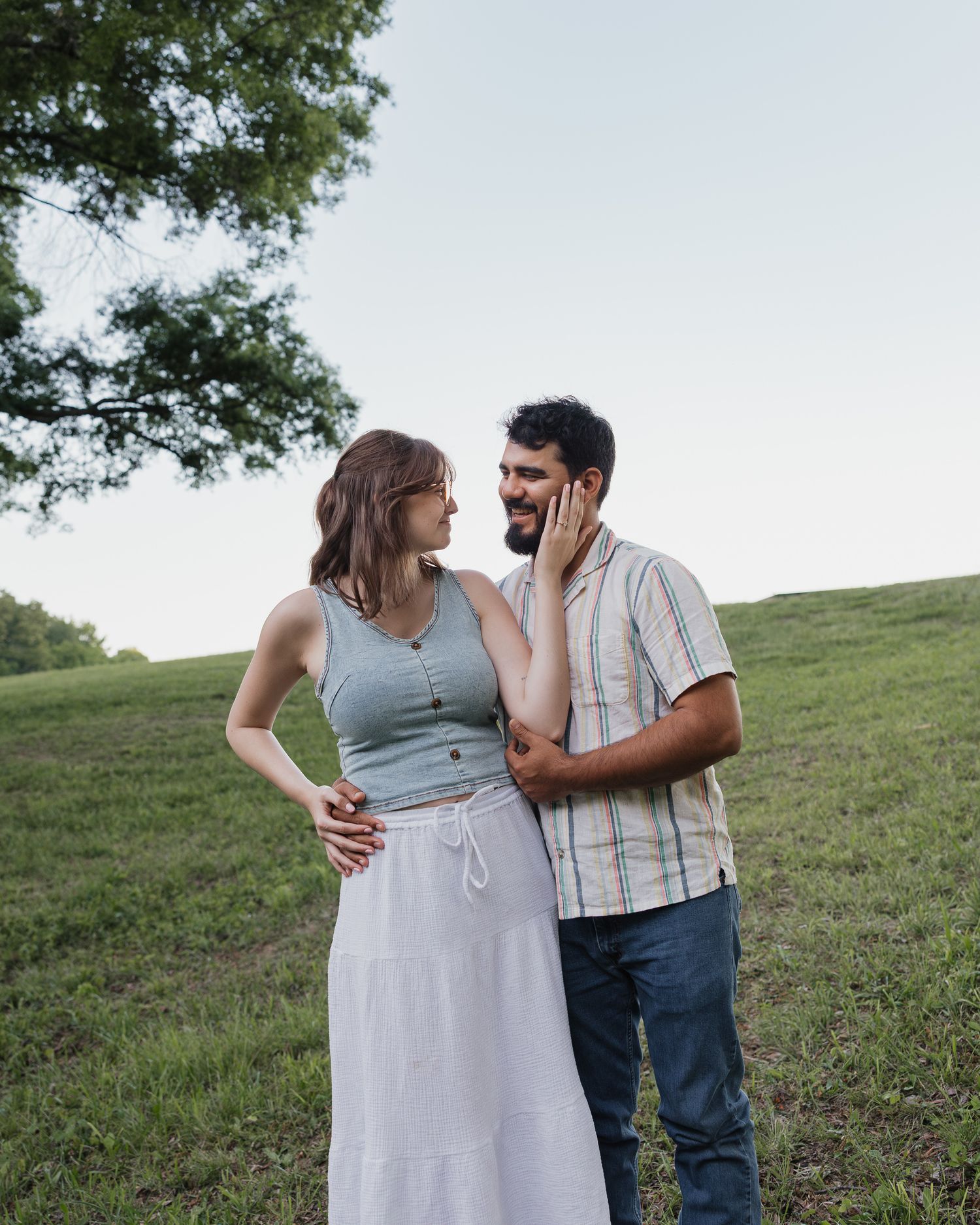 A couple shares a tender moment in a green meadow, woman in gray crop top and white skirt touching mans face.