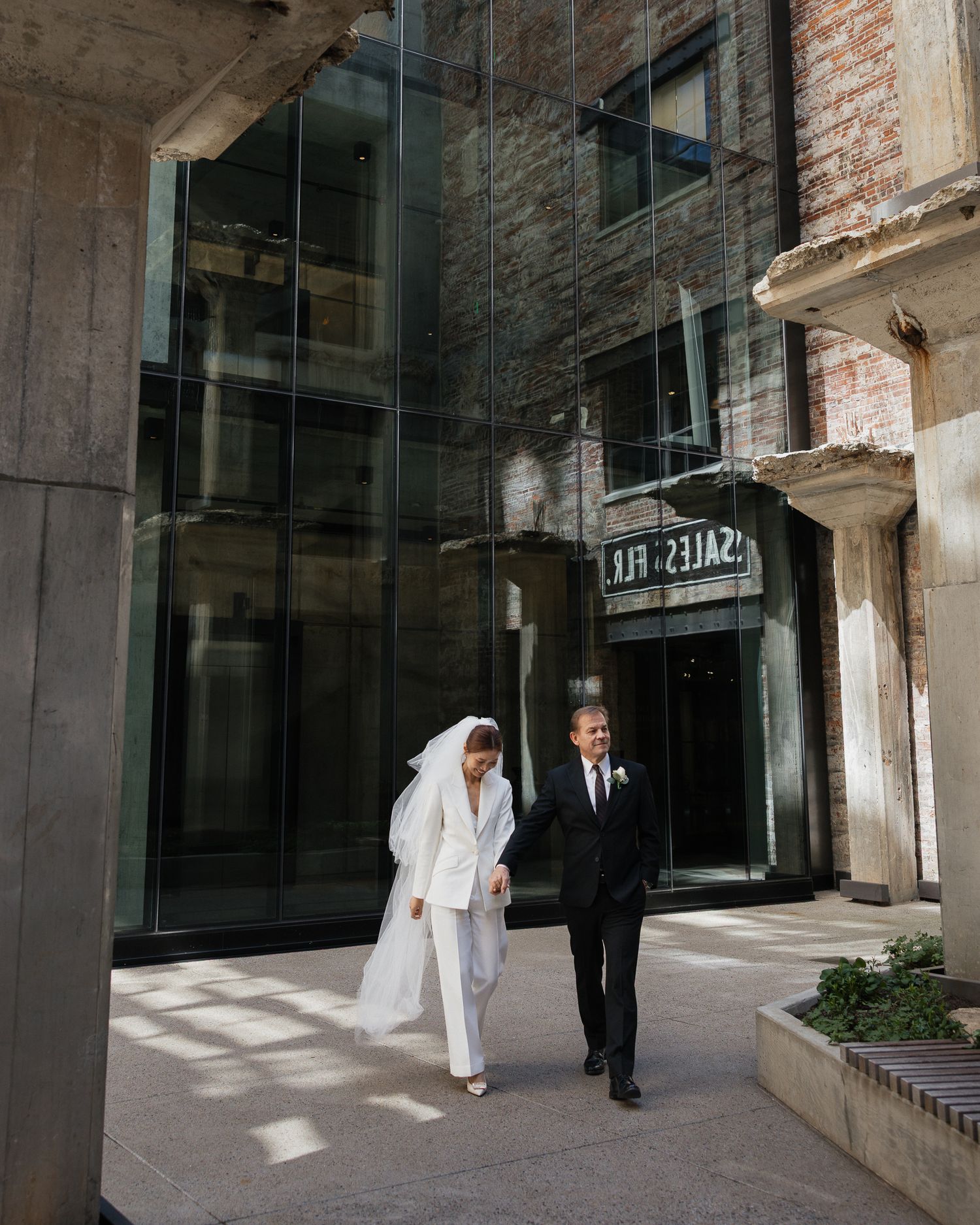 Bride in white pantsuit and veil walks with groom in black tuxedo outside a modern urban building in Downtown Nashville Tennessee.