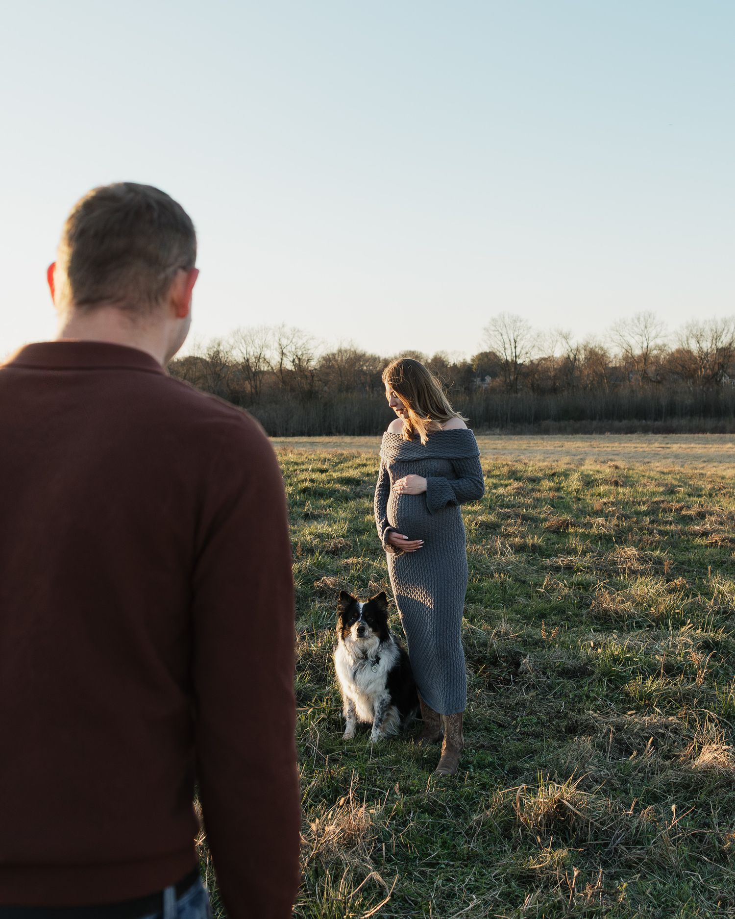 Couple embracing in an open field during sunset with their dog in the background.