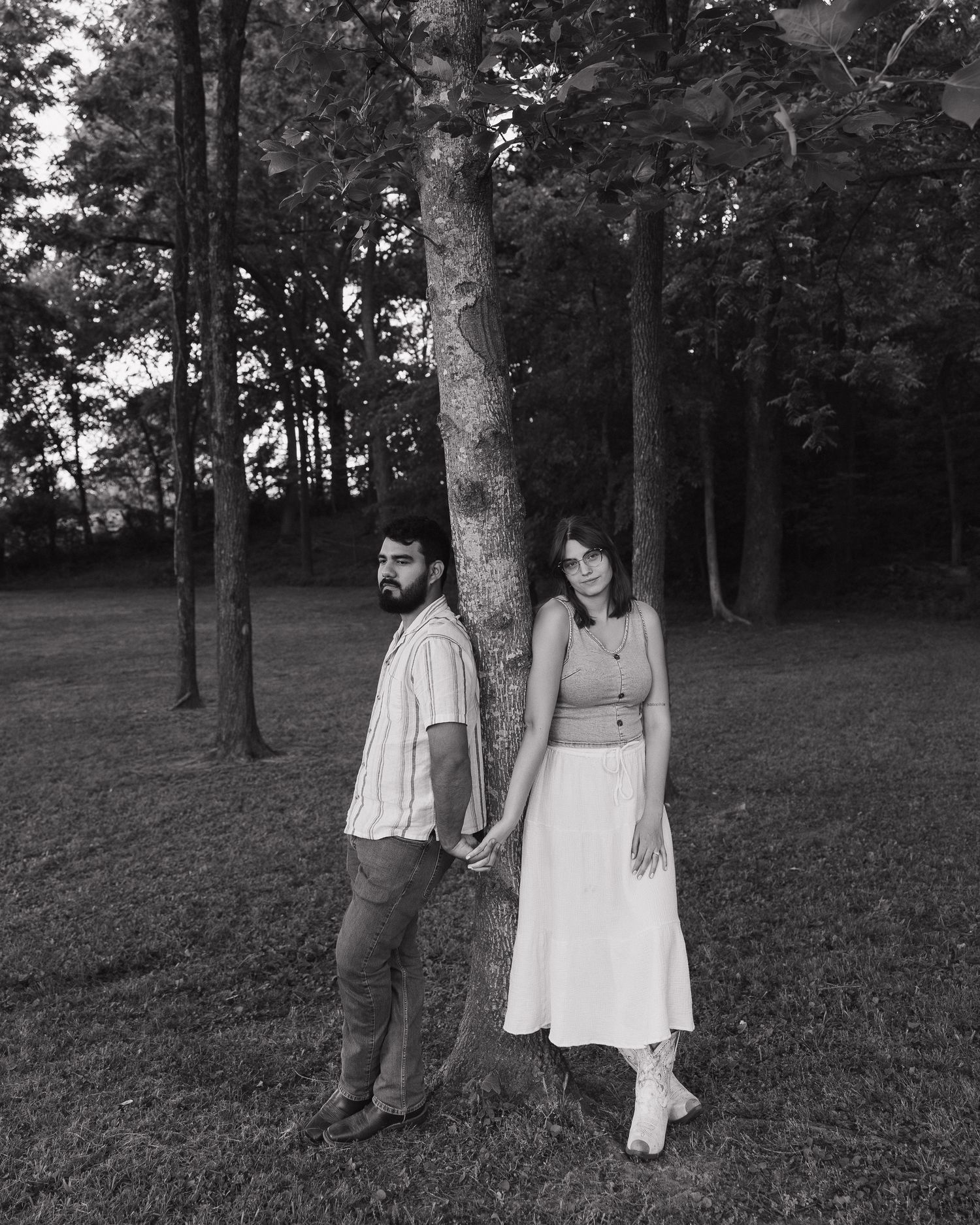 Black and white photo of a couple holding hands by a tree in a grassy park setting.